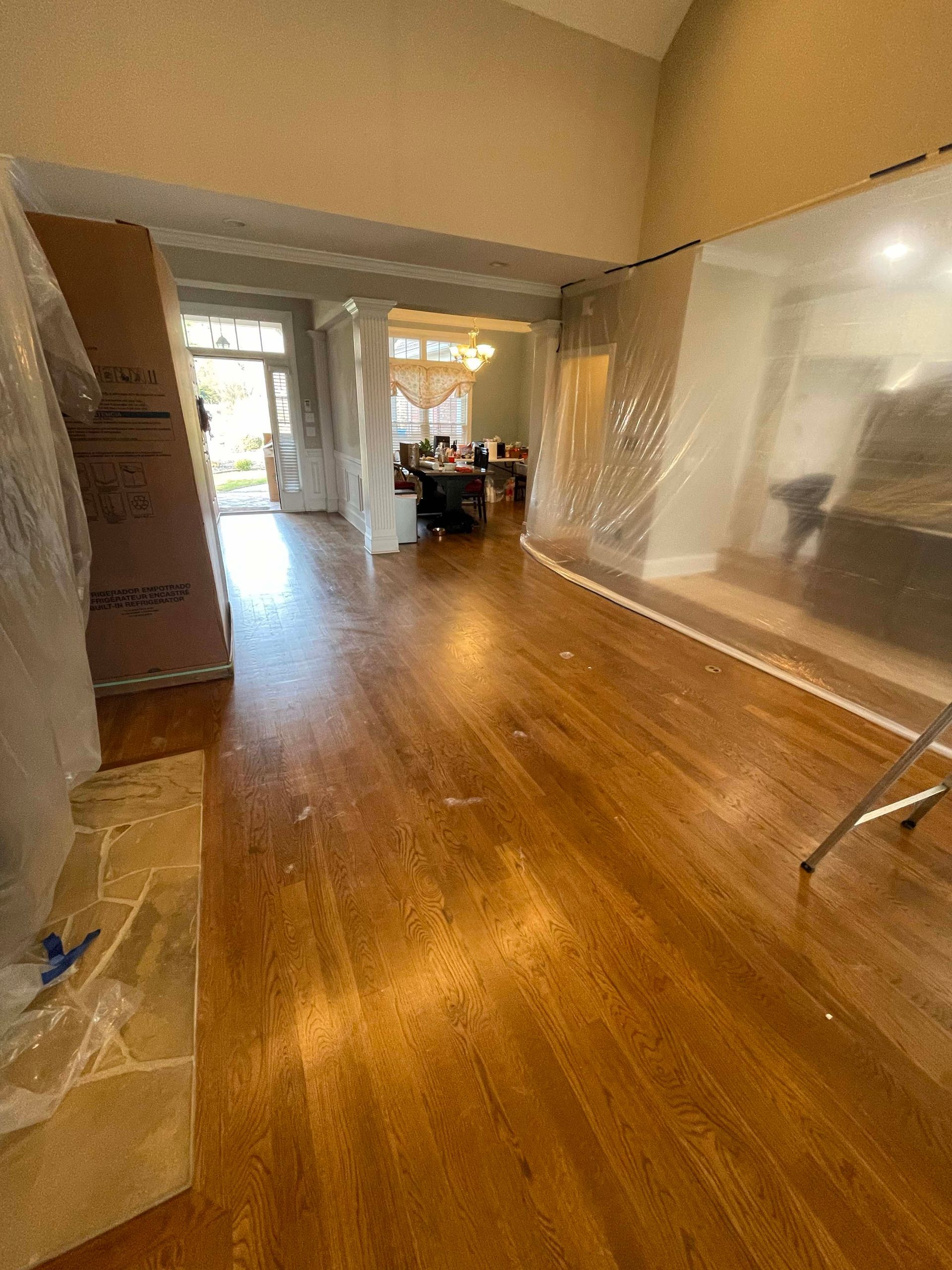 Hardwood floors in a home with plastic sheeting and doorway, leading to a dining area with a table and chairs.