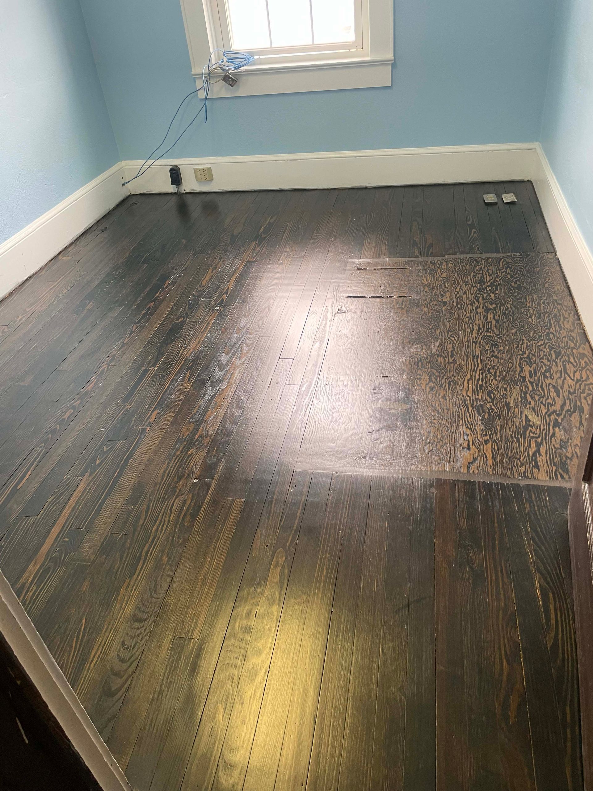 Dark, damaged hardwood floor in a room with blue walls, a white window, and trim.
