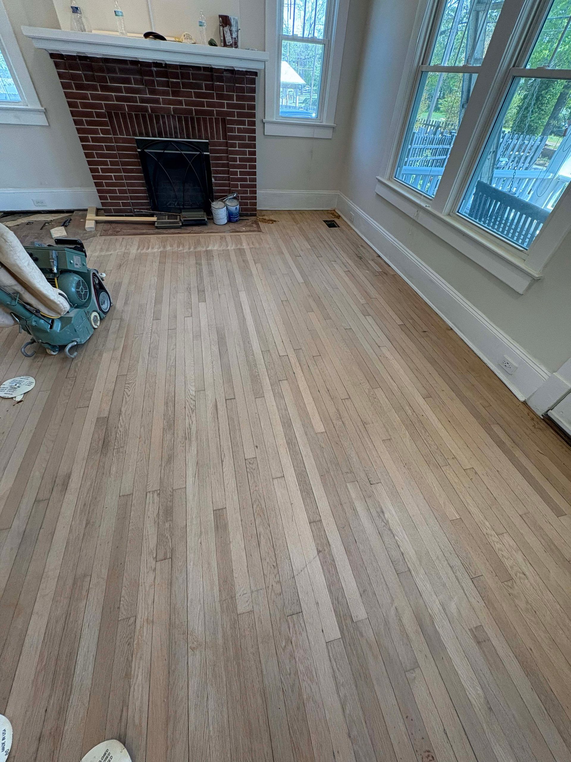 Hardwood floors in a room with a brick fireplace, near a window. A person is sanding the floor.