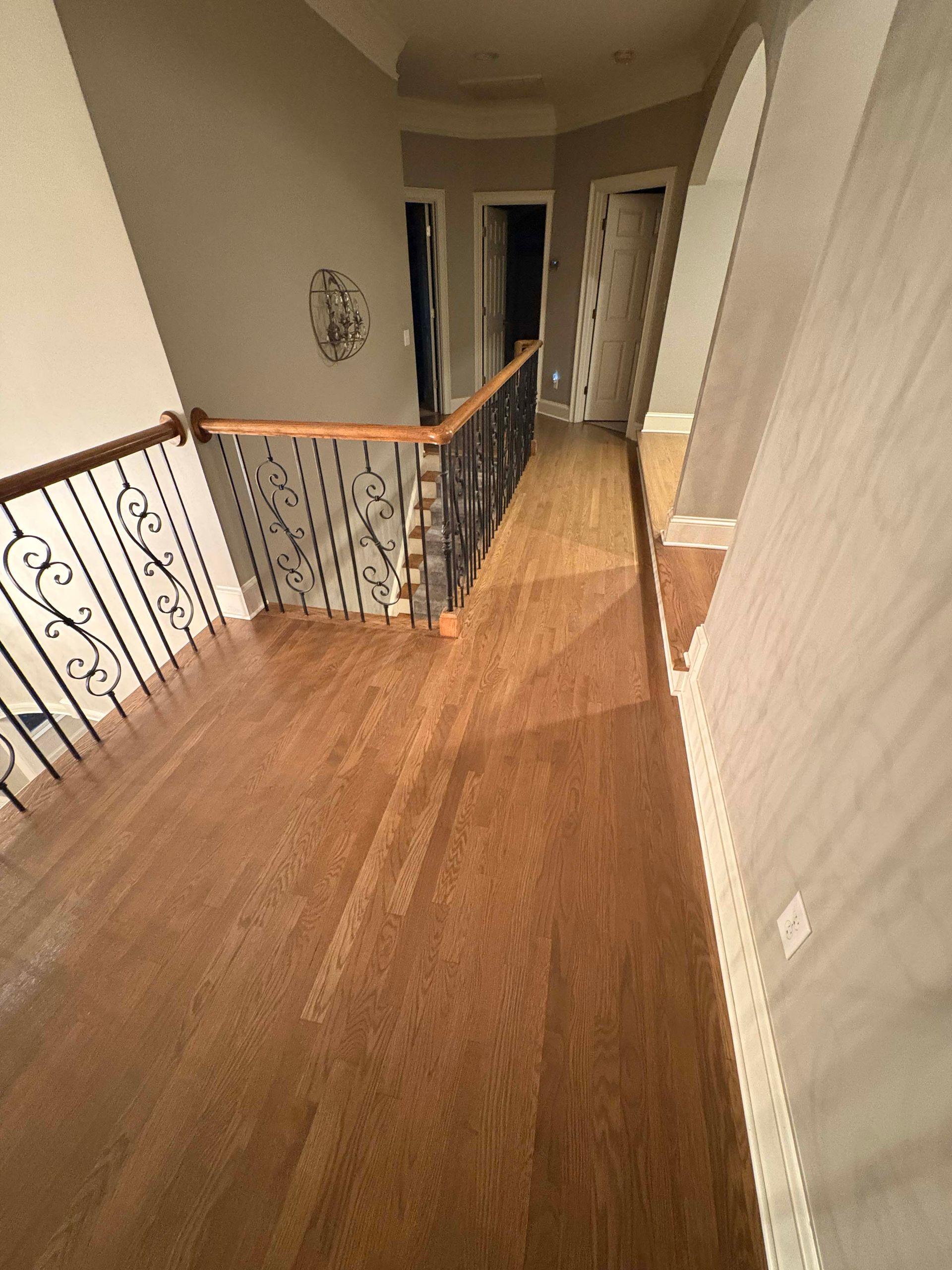 Wooden floor hallway with stairs and ornate iron railing, leading to doorways. Light and dark wood tones.