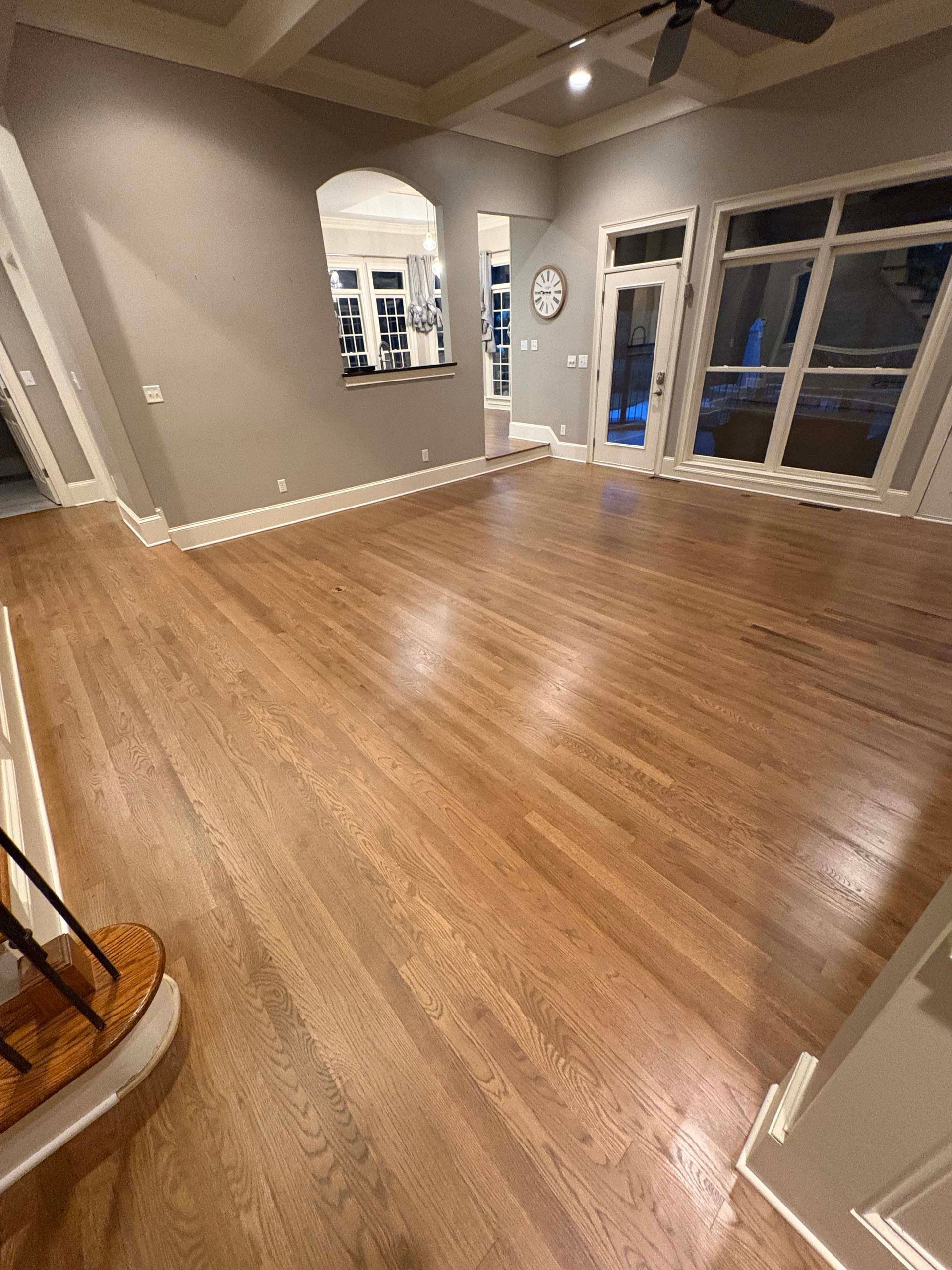 Empty living room with hardwood floors, gray walls, arched window, and large windows with outdoor view.