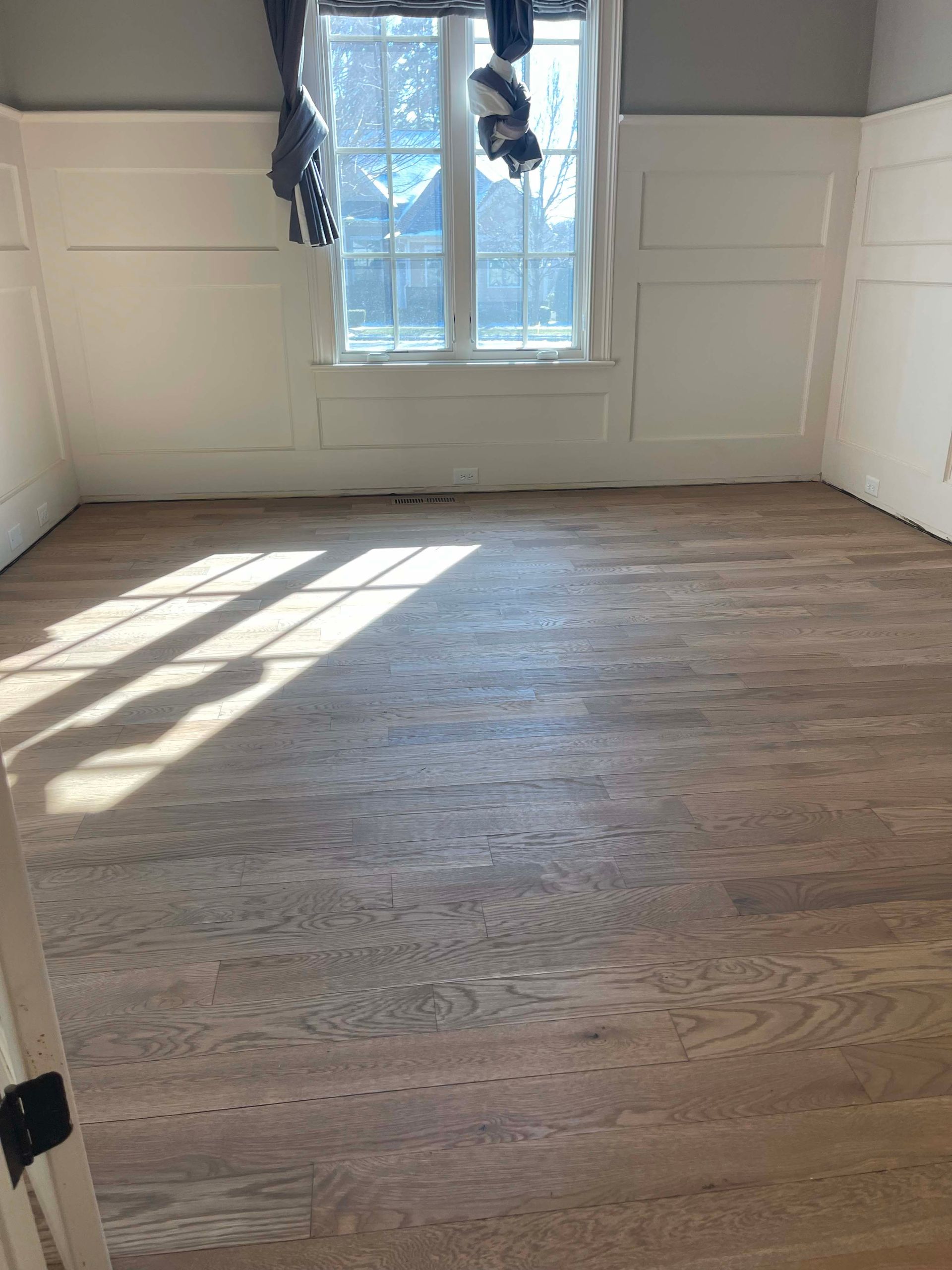 Empty room with light wood floors and white paneled walls, sunlit window.