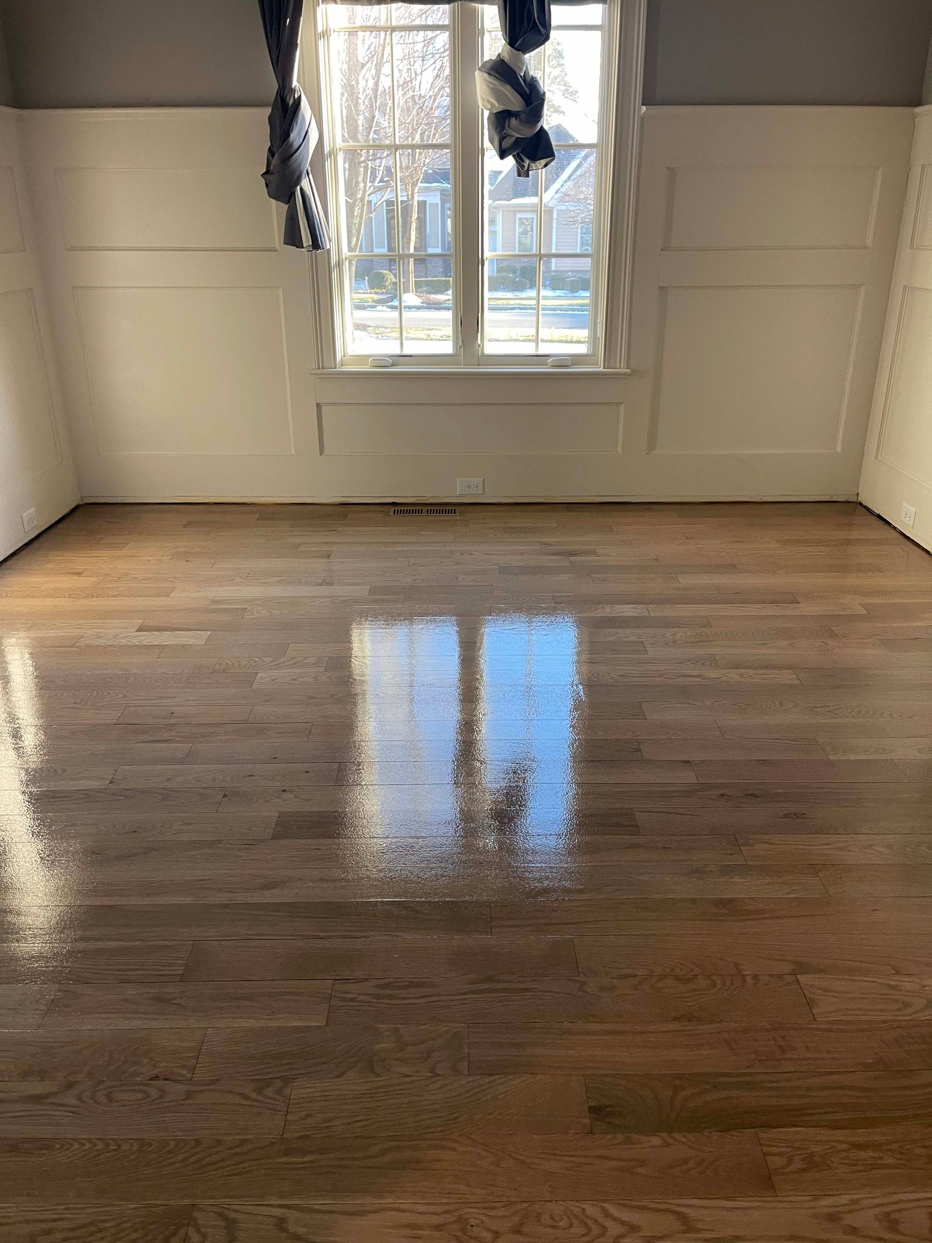 Reflective, newly finished wood floor in a room with white paneled walls and a window.