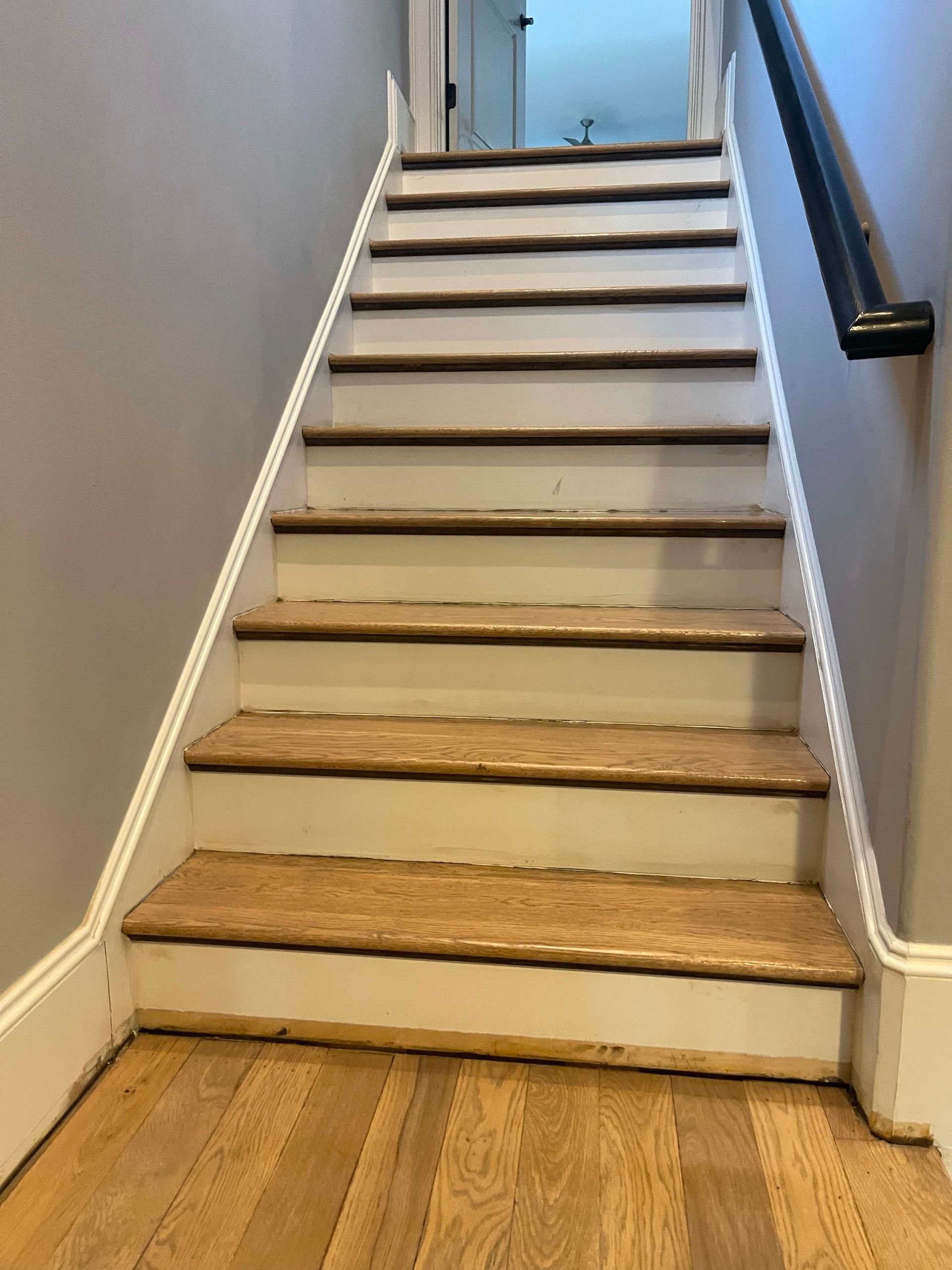 Staircase with wooden steps and white risers, leading upwards. A black handrail is on the right.