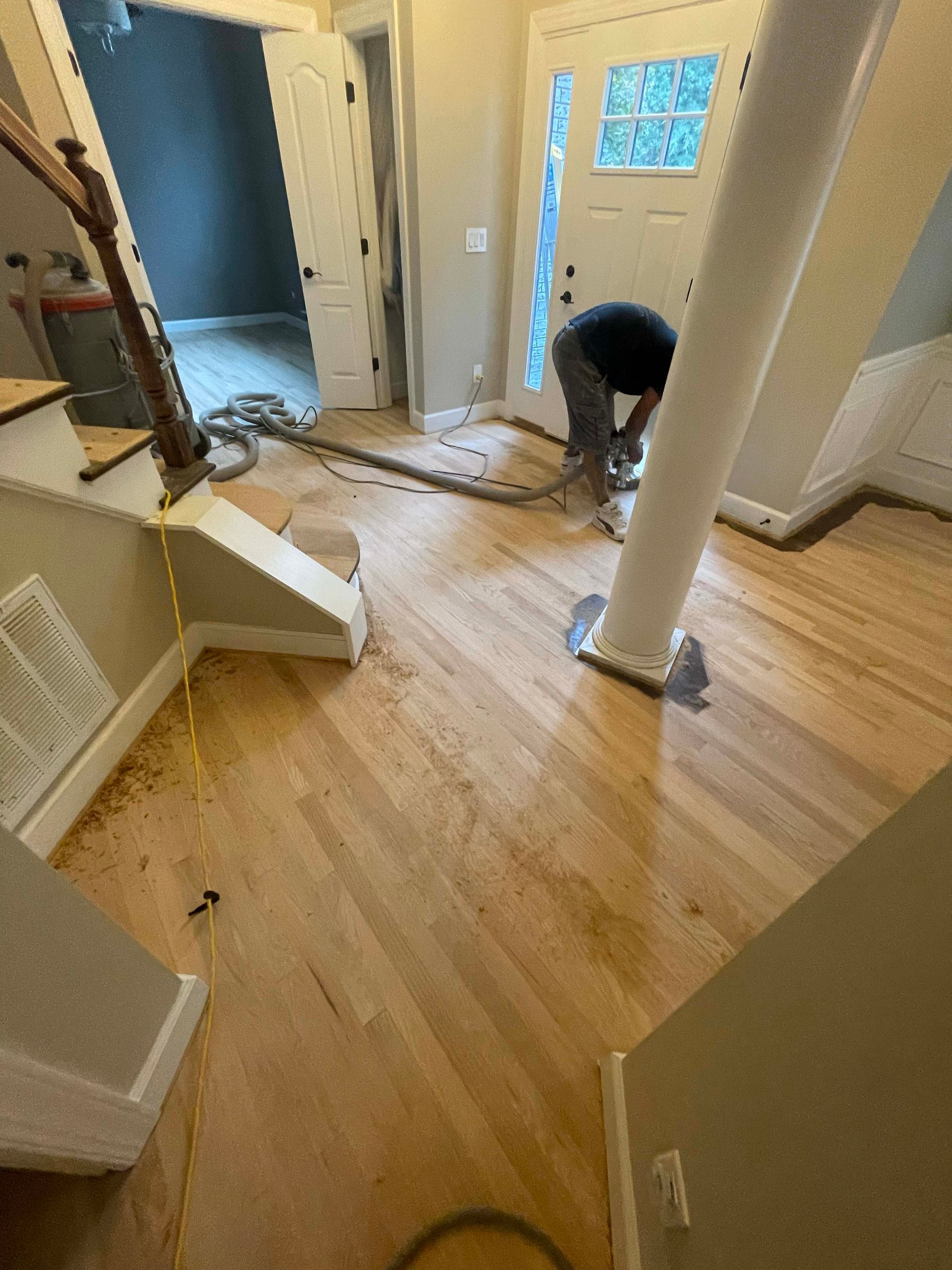 Man sanding hardwood floor near a white support column and stairs. Dust covers the wood.