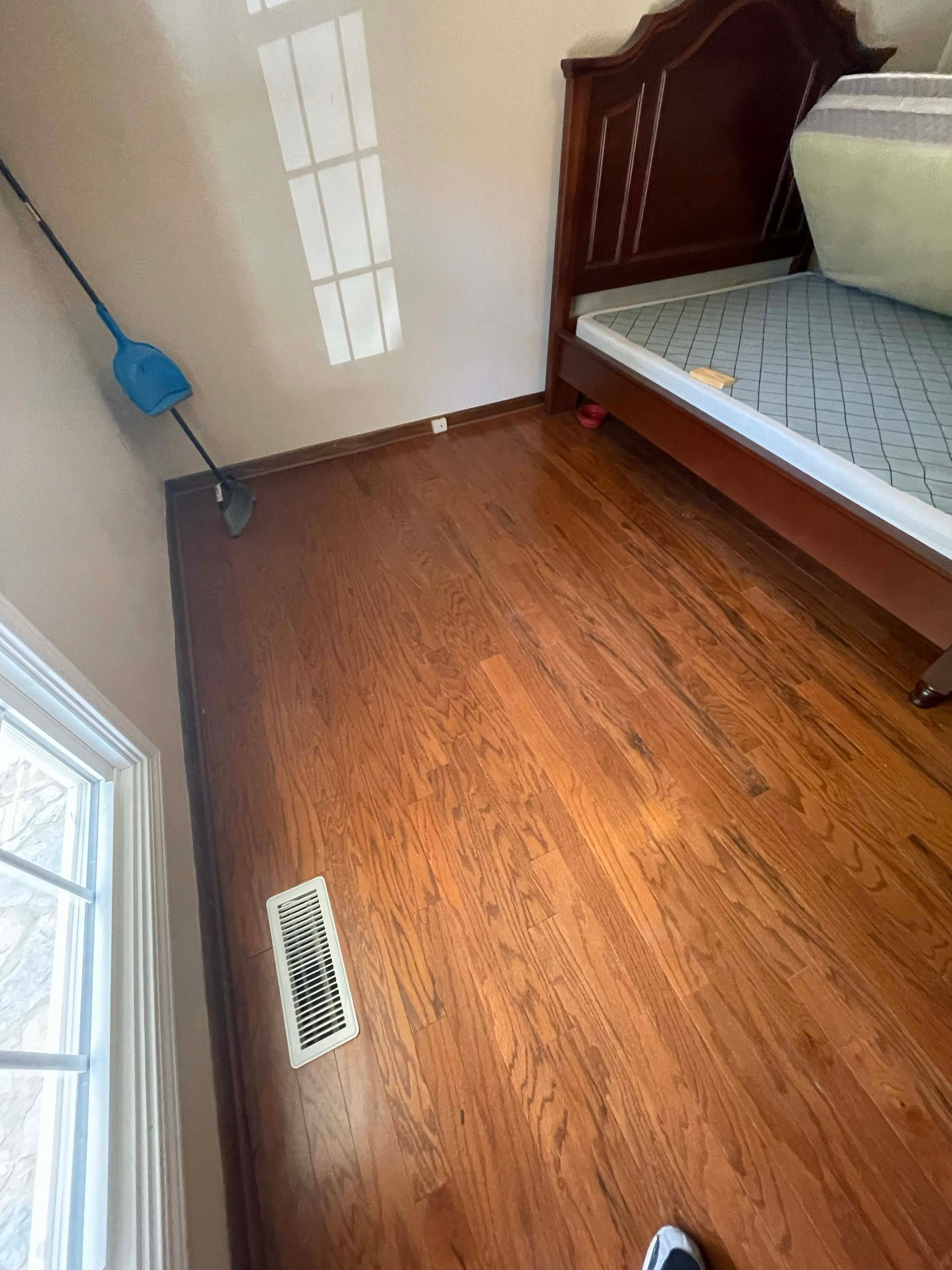 Wooden bedroom floor with bed, vent, and broom.