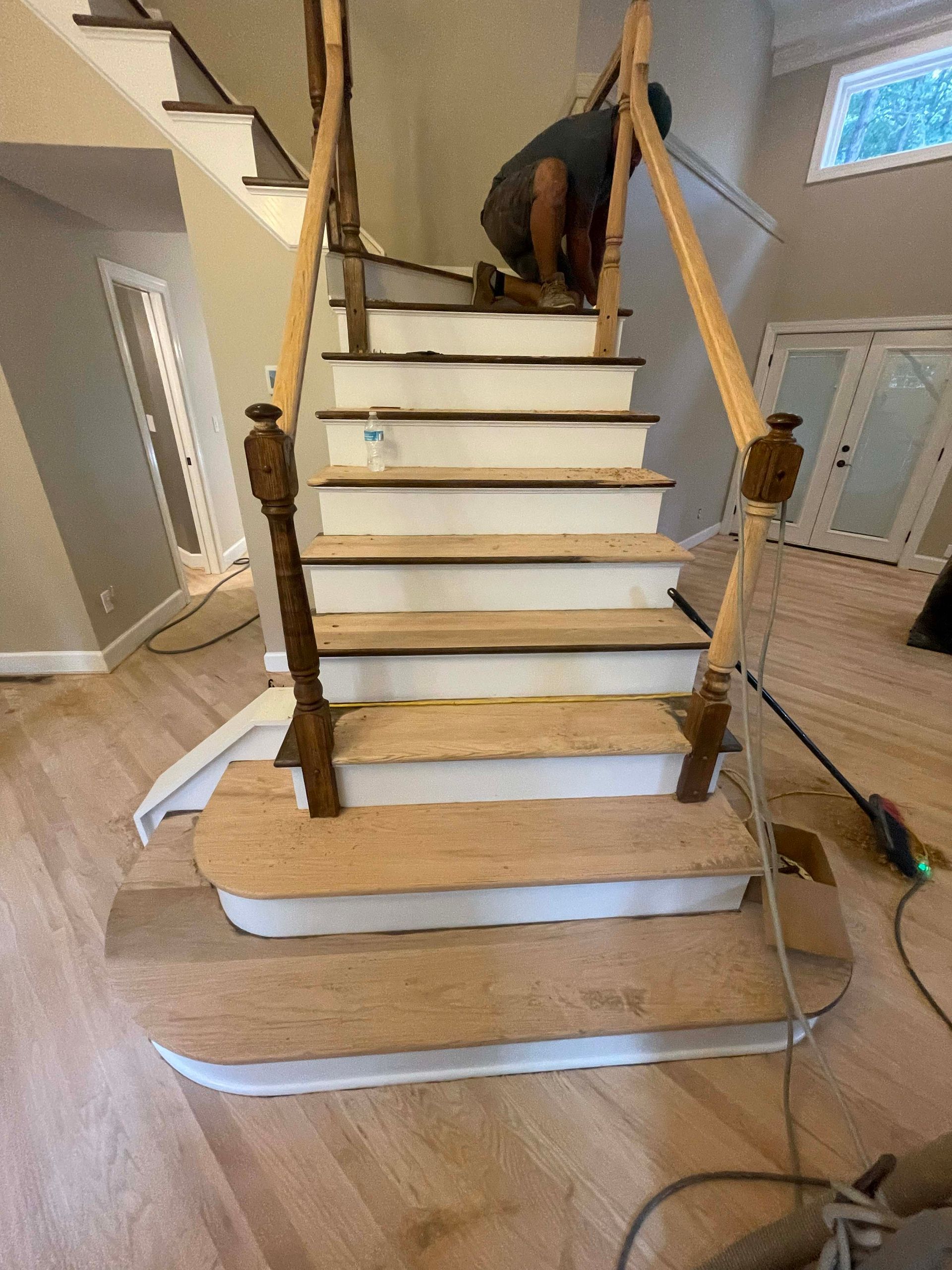 Person installing wood treads on a staircase with white risers and wooden banisters.