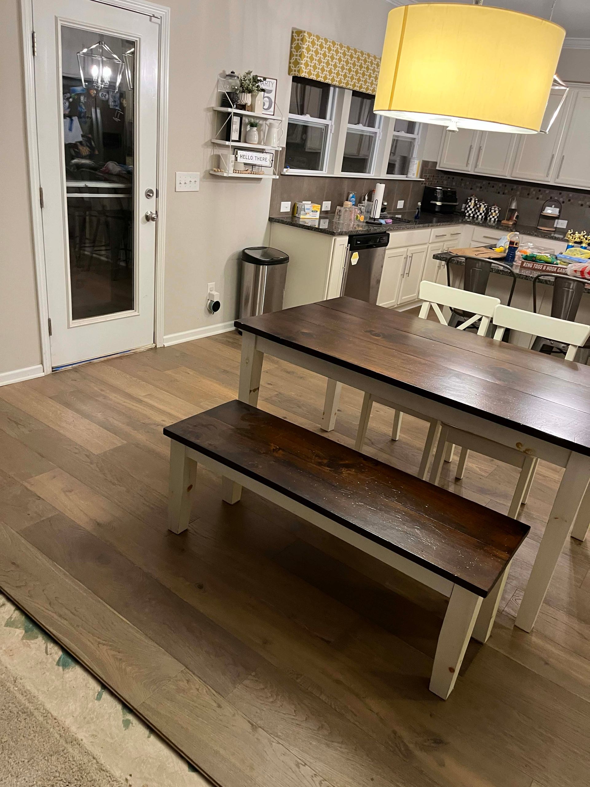 A dining area with a wooden table and bench, white trim, and a kitchen in the background.