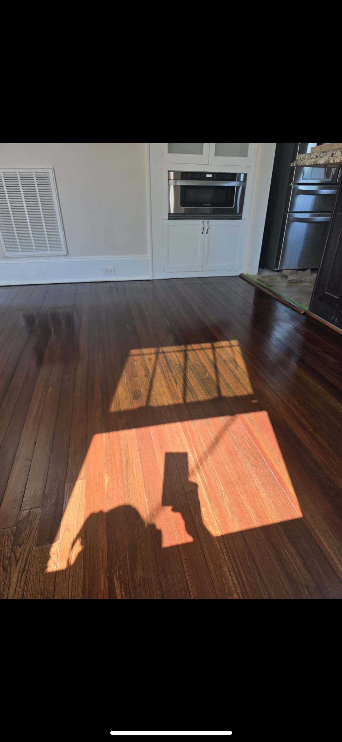 Shadow of a person holding a phone on wooden floor; kitchen in background with vent and built-in microwave.
