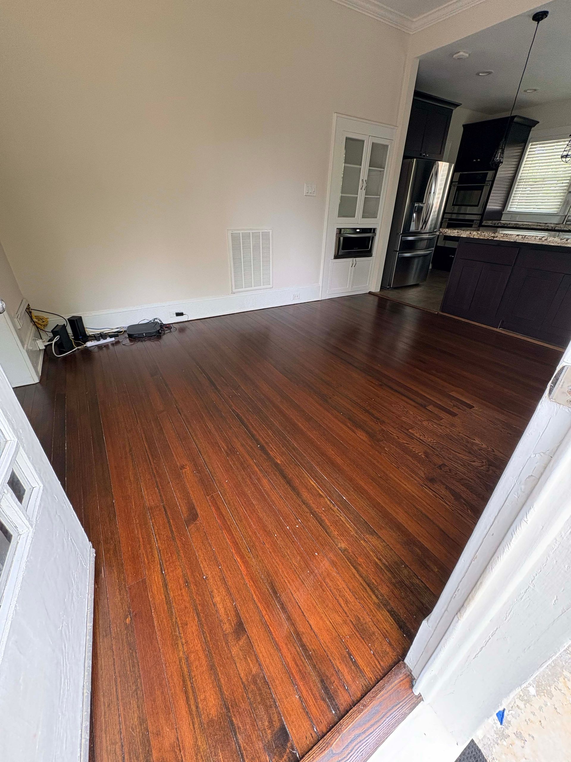 Dark wood floor in a room, leading to a kitchen. Light-colored walls and a white door frame.