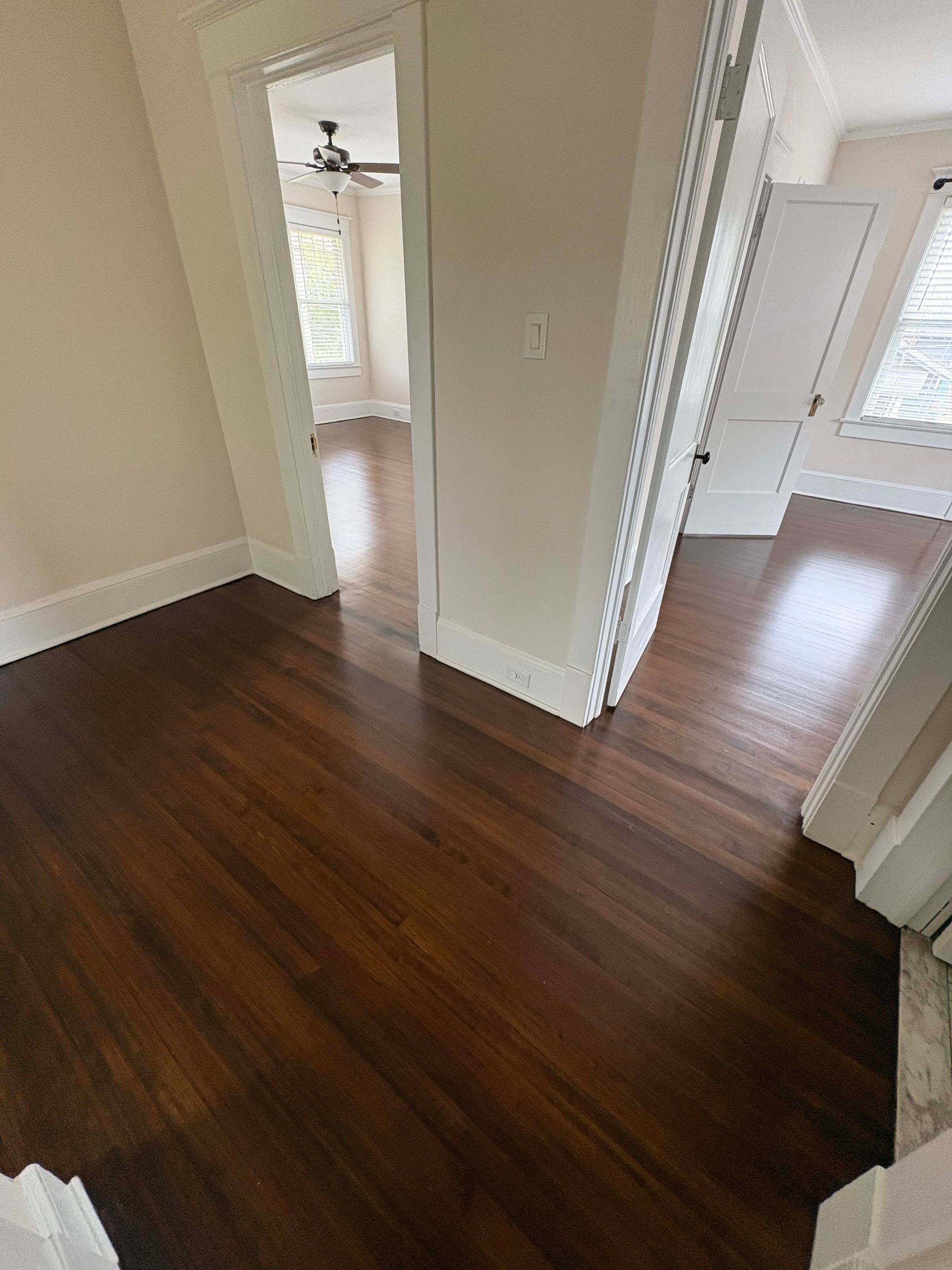 Dark wooden floors in a room with white walls, leading to other rooms, with a ceiling fan visible.