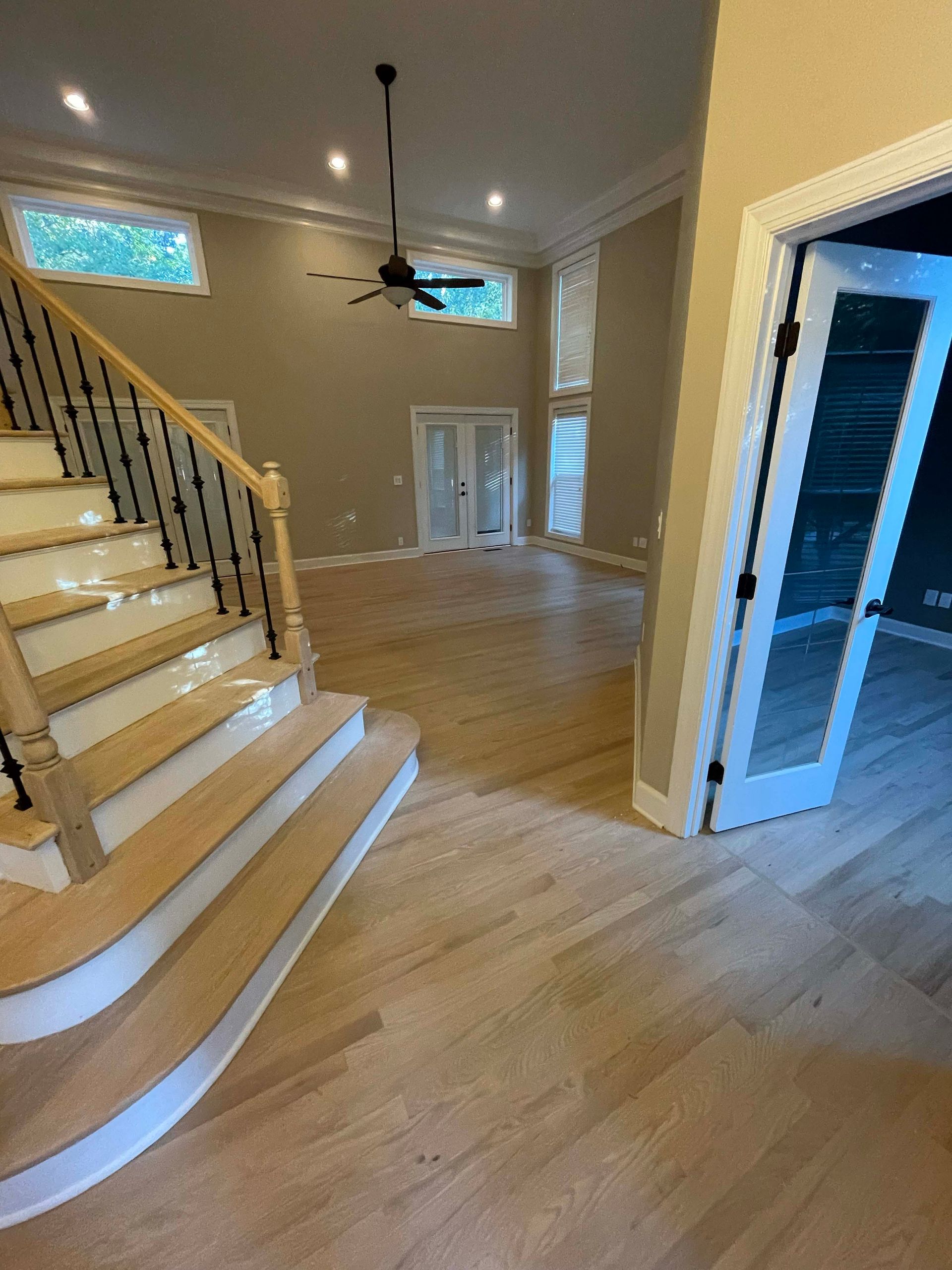 Empty, two-story room with hardwood floors, staircase, and double doors. Beige walls, natural light.