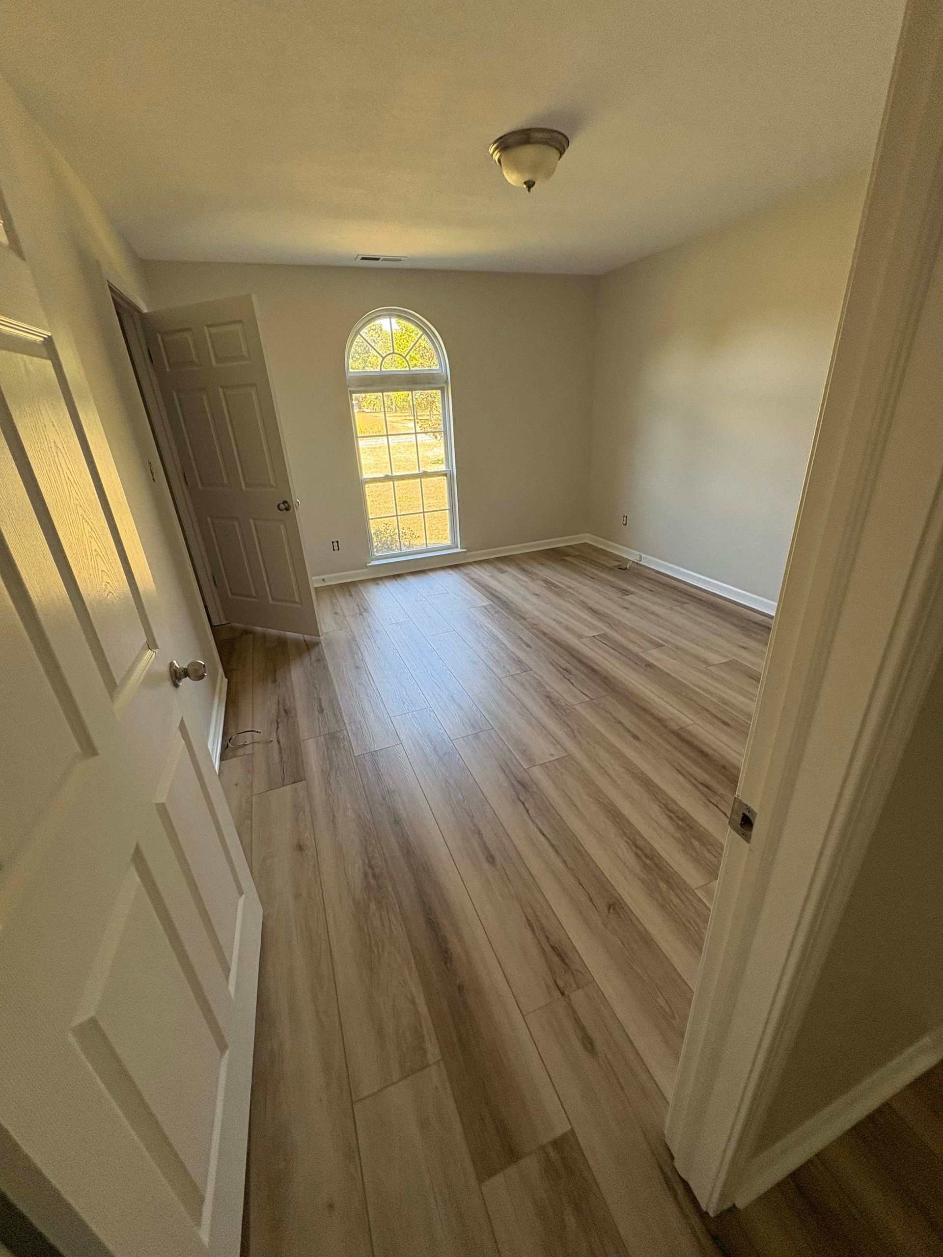 Empty room with wooden floors and arched window. Light walls, two doors, and a ceiling light.