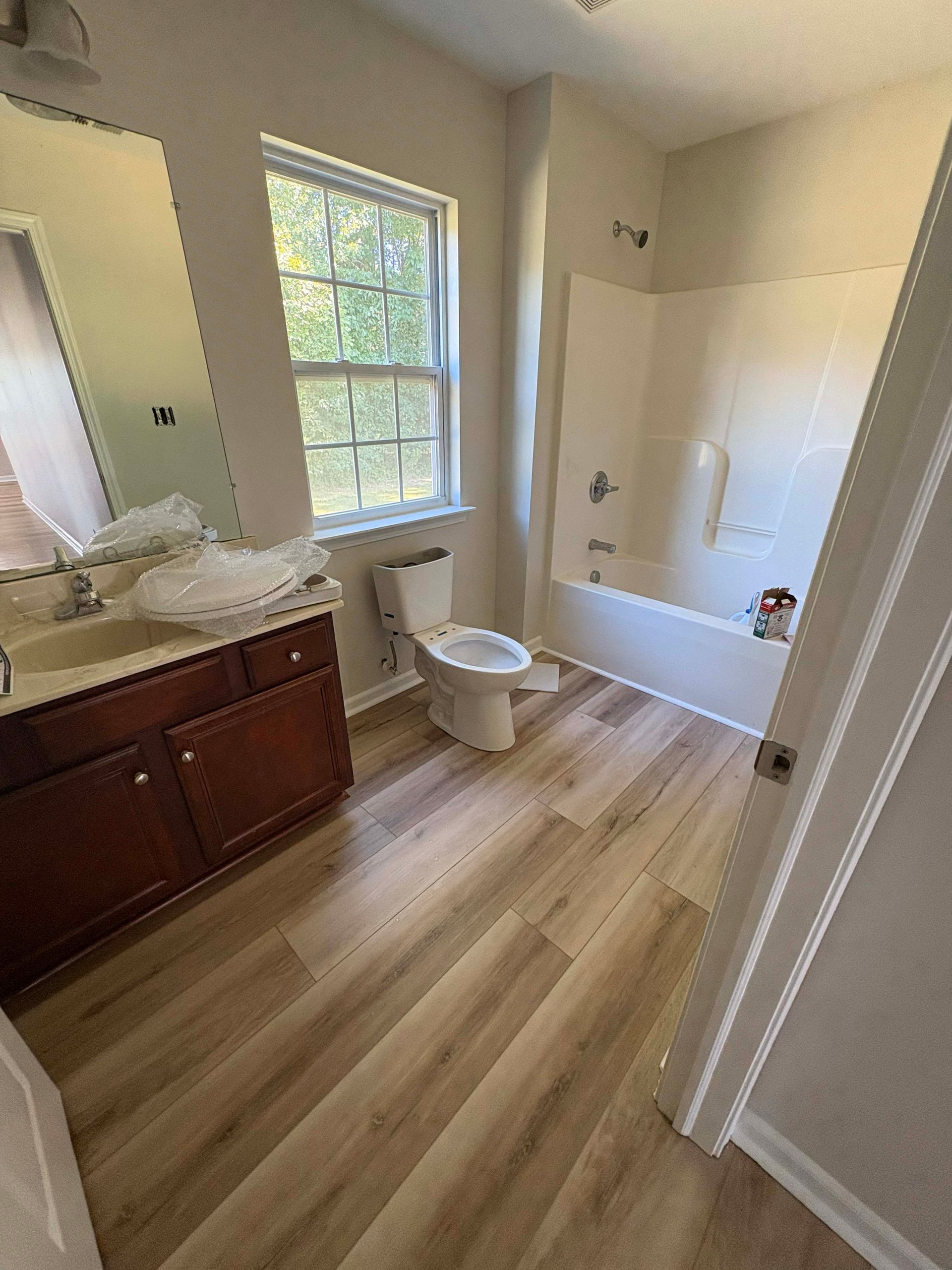Bathroom with wood-look flooring, dark wood vanity, toilet, tub/shower, and a window.