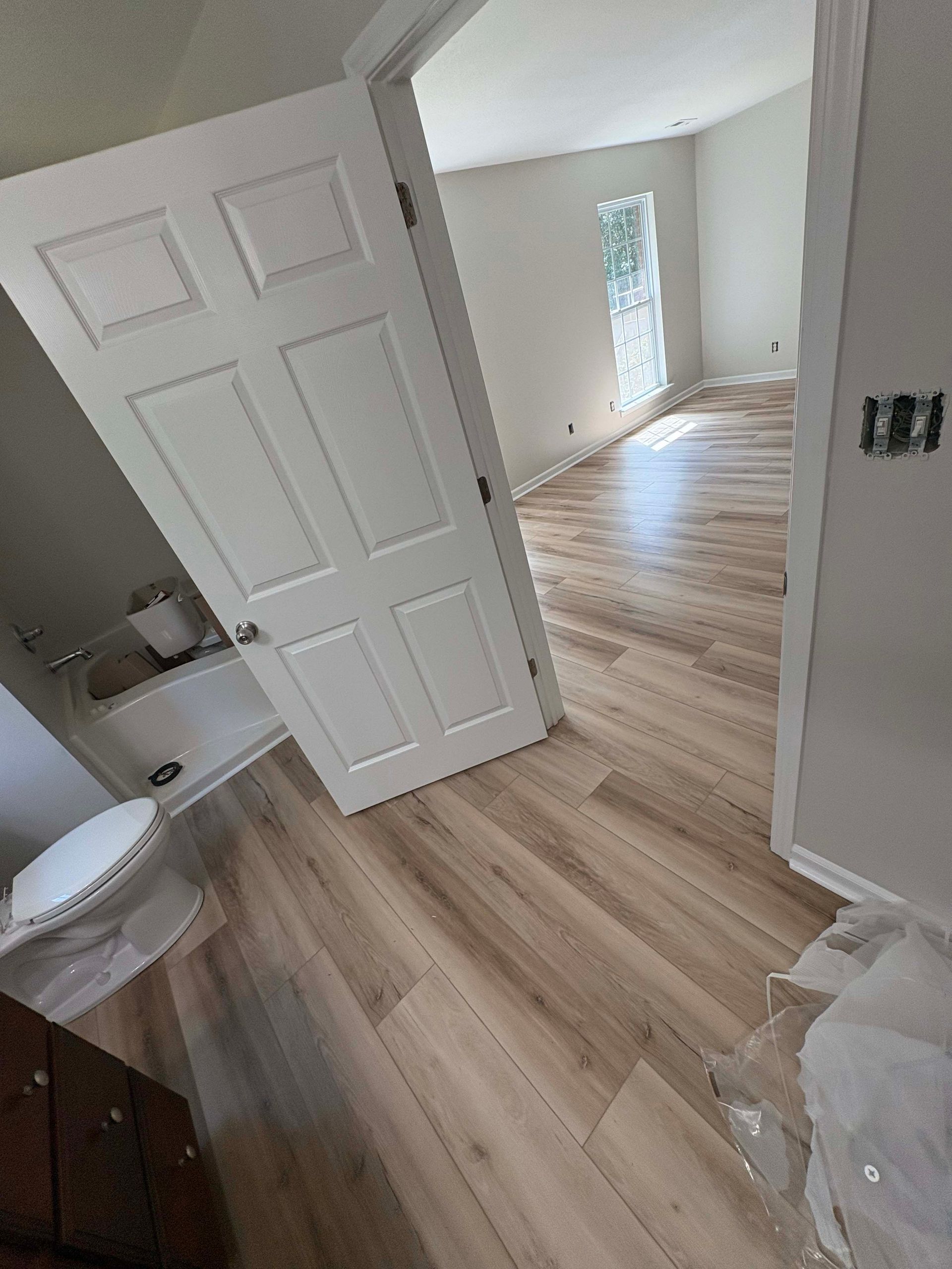 Bathroom and bedroom with light wood-look flooring and white walls. Open door reveals toilet and bathtub.