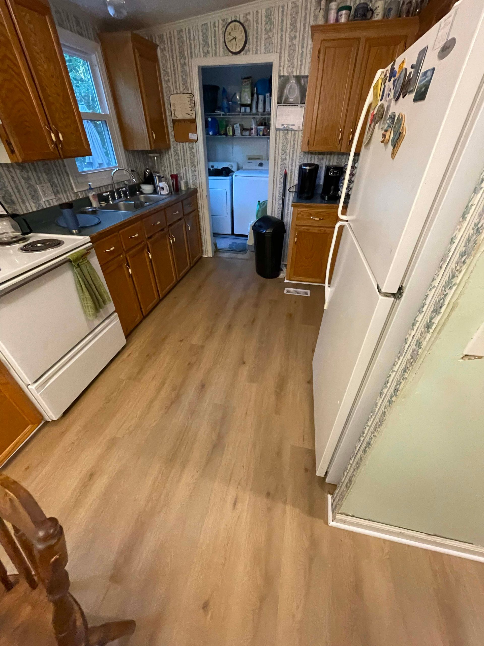 A kitchen with wood cabinets, white appliances, and a light wood floor. A doorway leads to a laundry area.