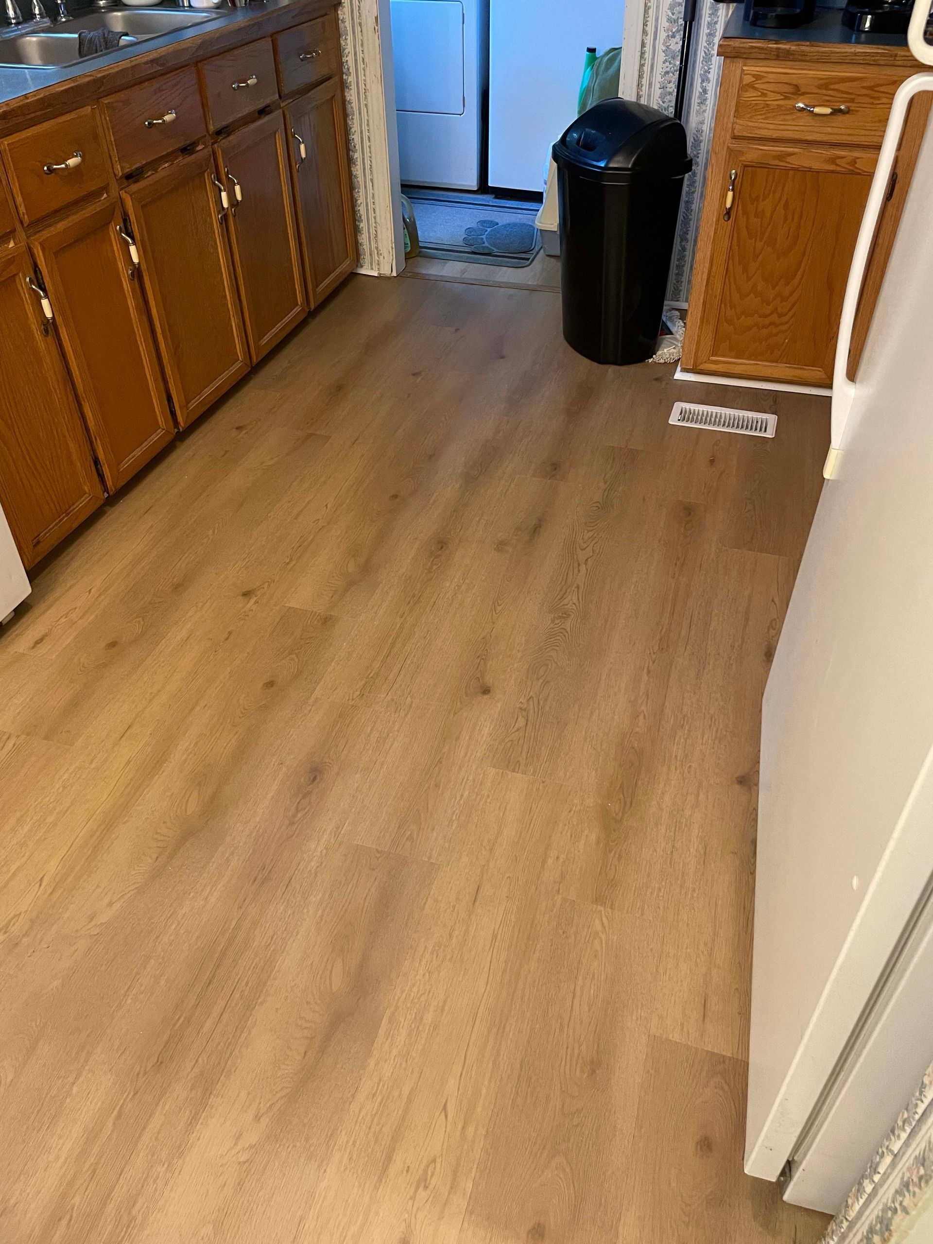 Kitchen with wood-look flooring and brown cabinets. A black trash can is next to a refrigerator.