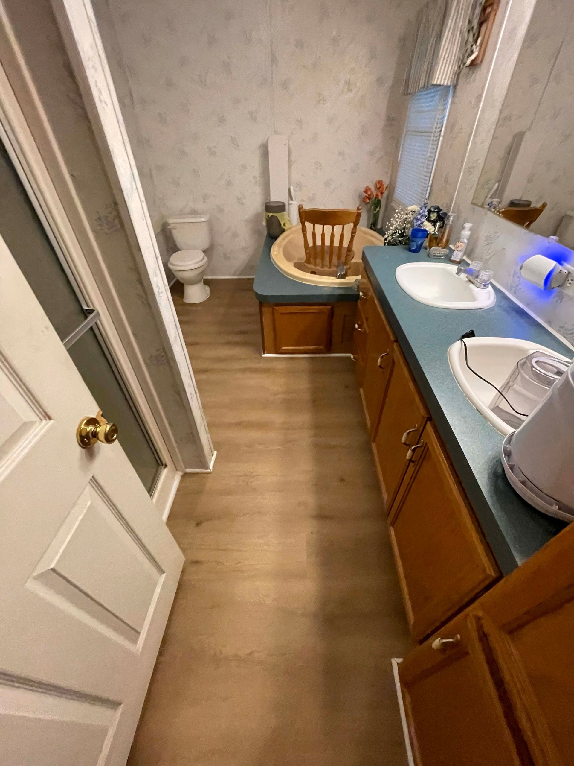 Bathroom with toilet, vanity with two sinks, and wooden chair on a cabinet. Light blue countertops, wooden cabinets.
