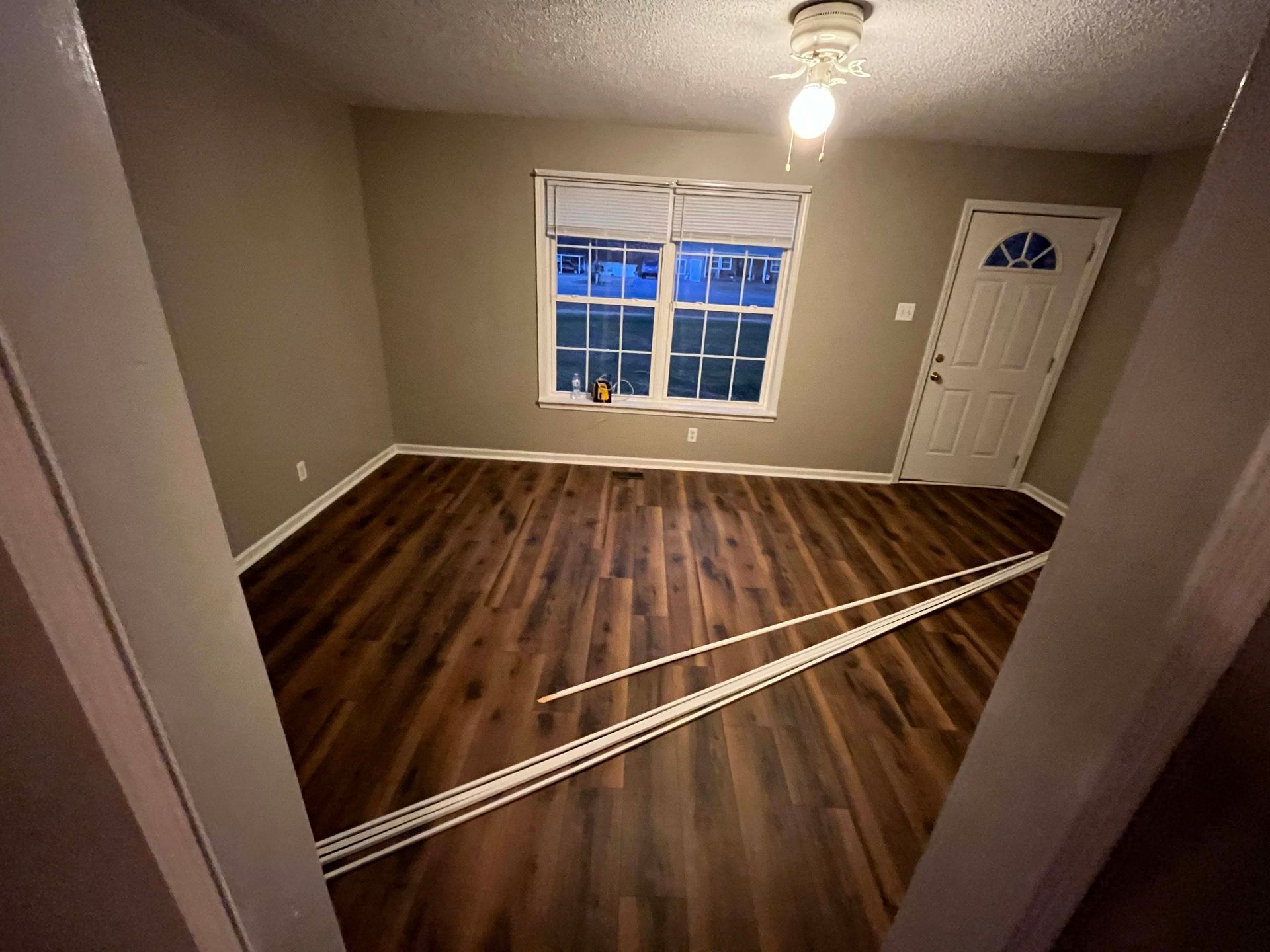 Empty room with dark wood-look flooring, tan walls, window with blinds, and a white door.