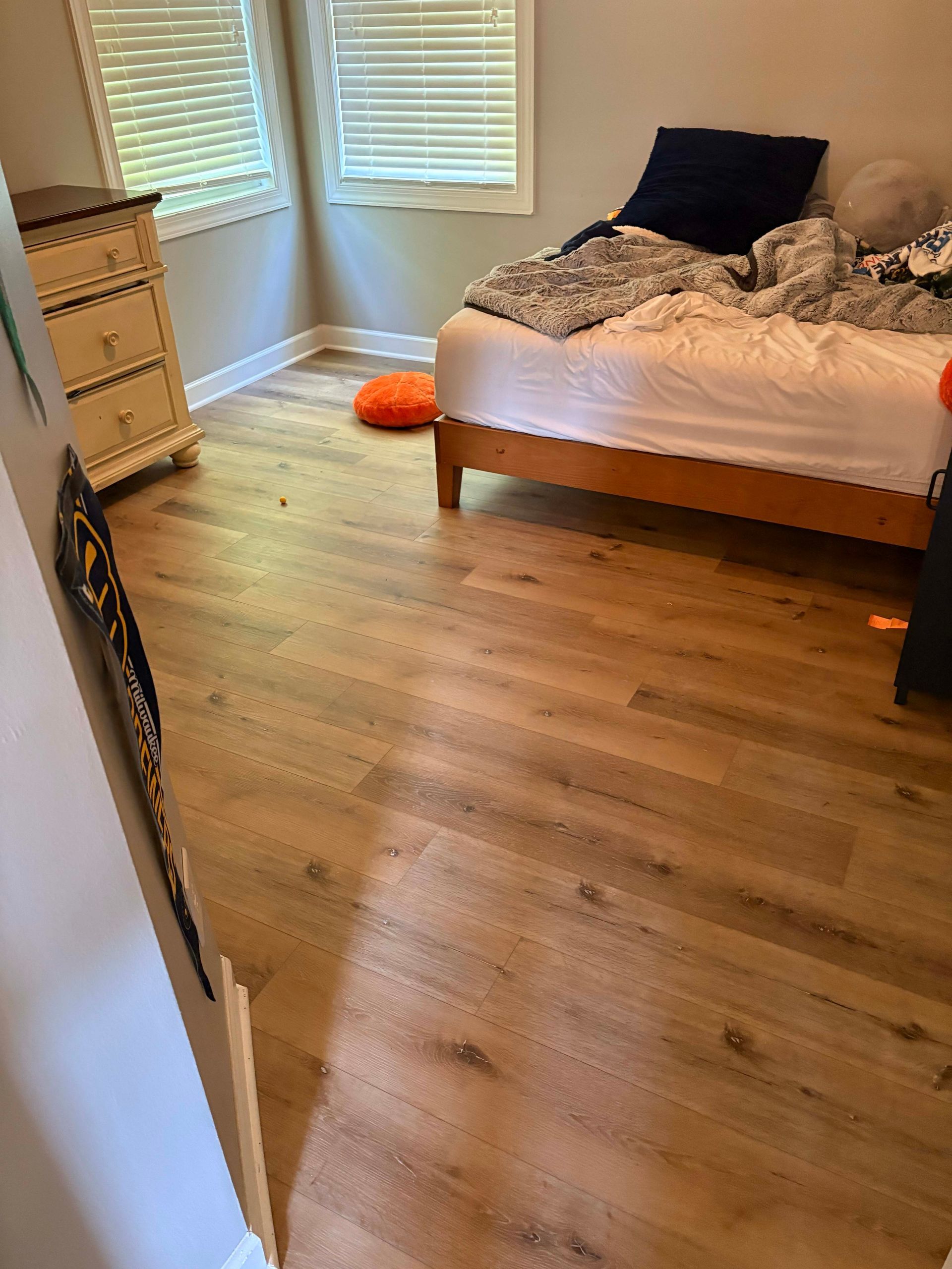 Bedroom with wood-look flooring, a bed with rumpled covers, and a dresser. Windows and an orange ball are also visible.