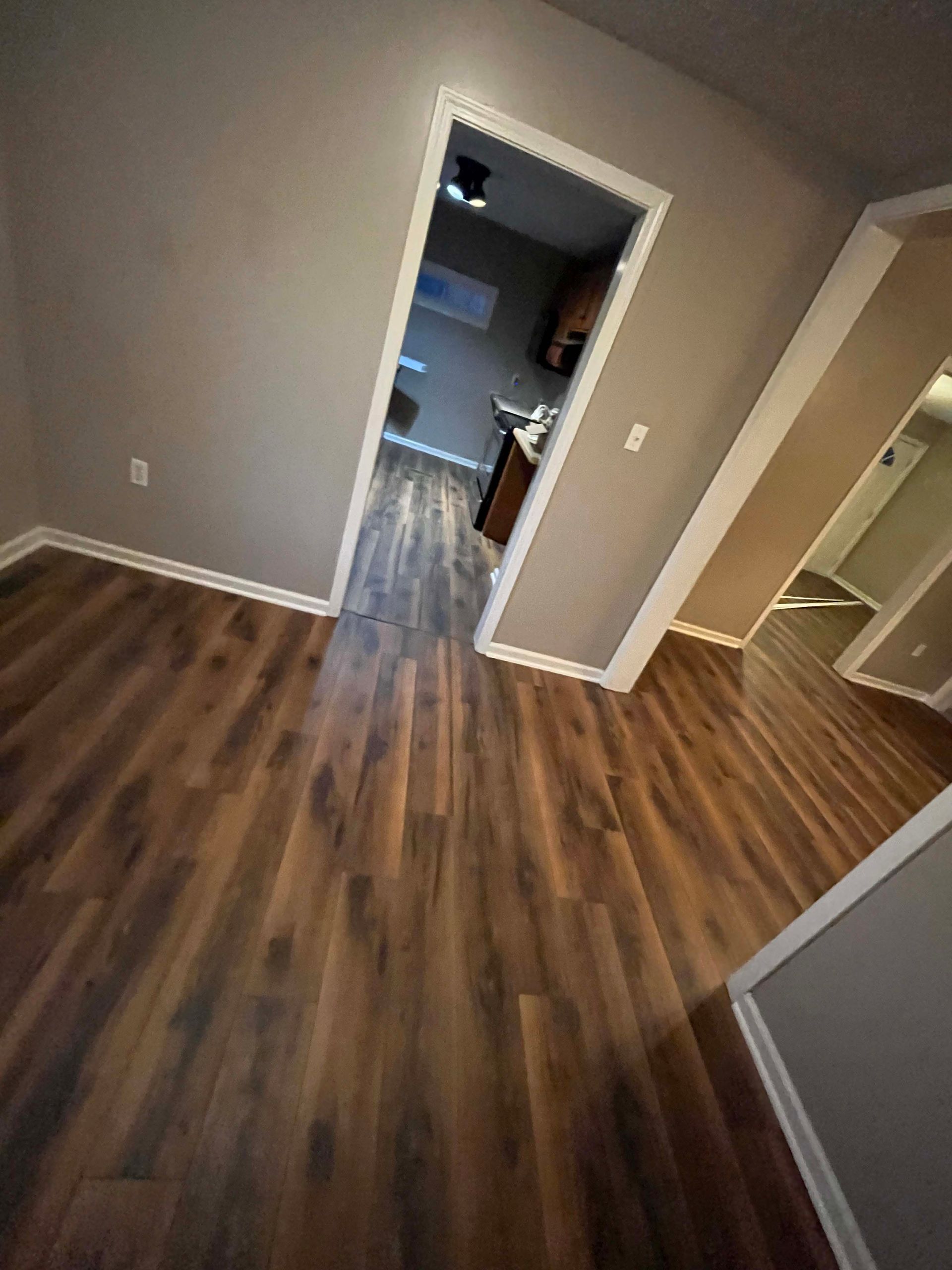 Room with dark wood flooring, tan walls, and a doorway leading to a kitchen with cabinets.