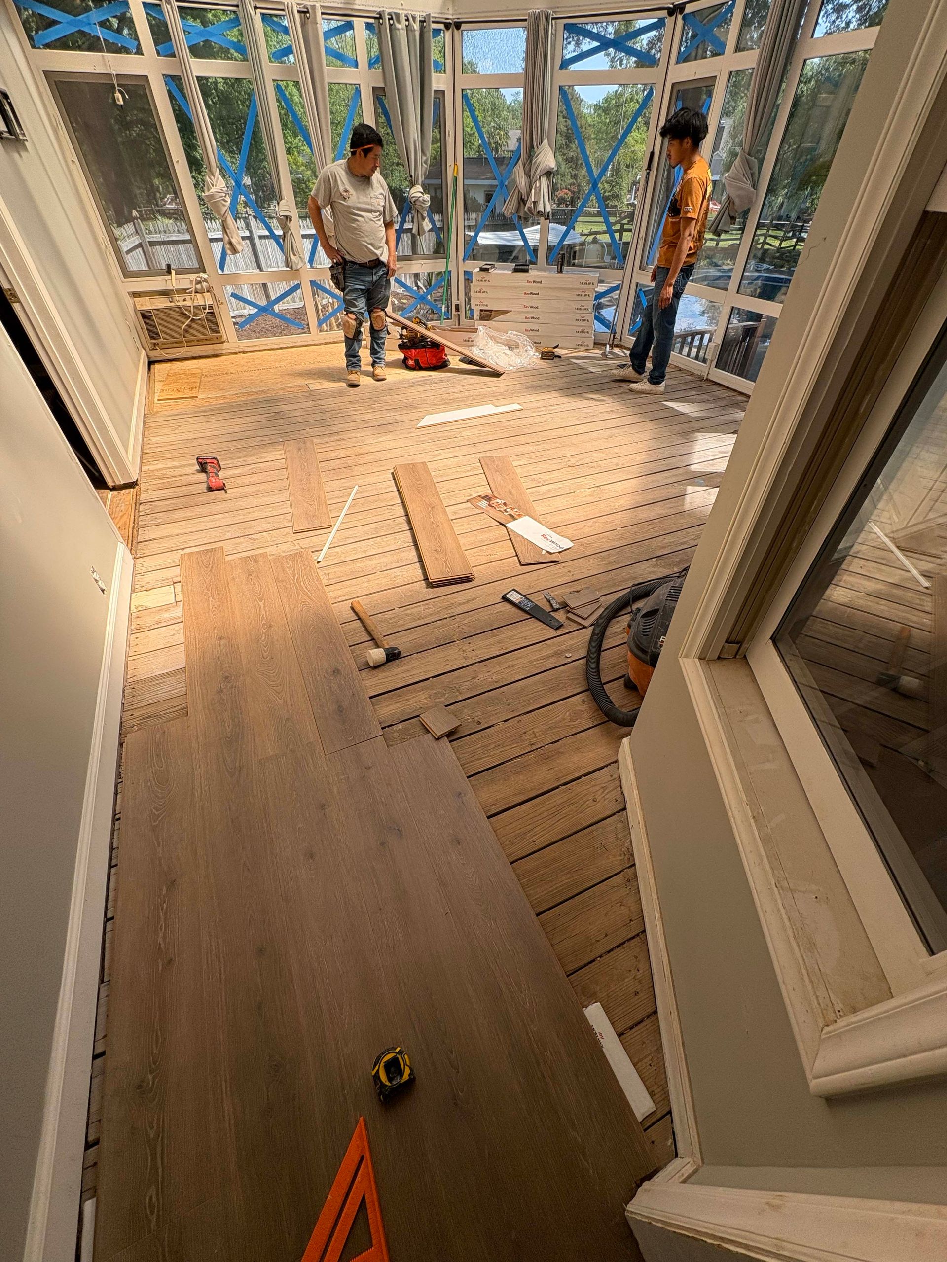Two workers installing wood flooring in a sunroom with large windows.