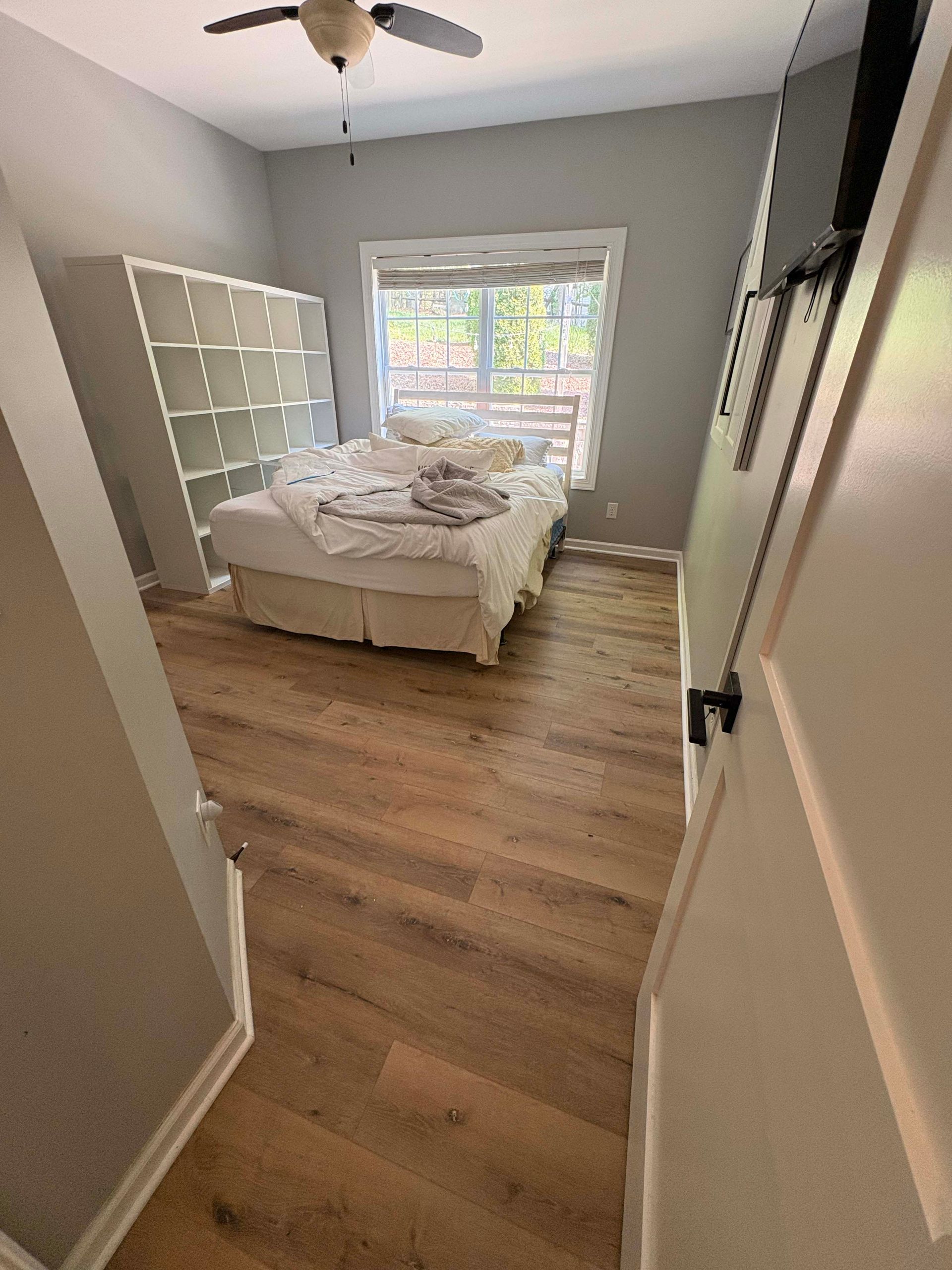 Bedroom with bed, open shelving unit, window, and wood-look flooring.