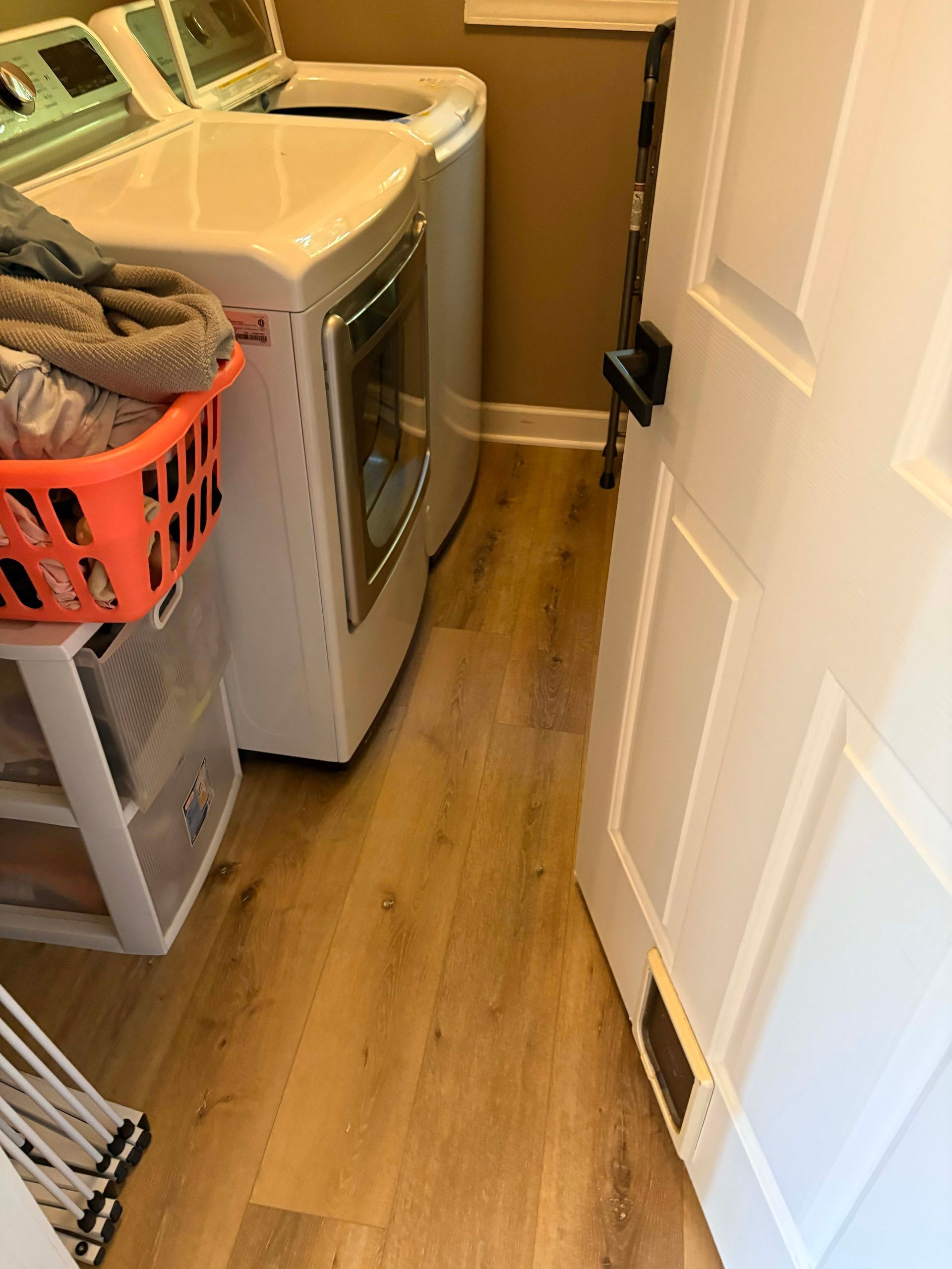 Laundry room with washer, dryer, orange basket of clothes, and open white door. Wood-look flooring.