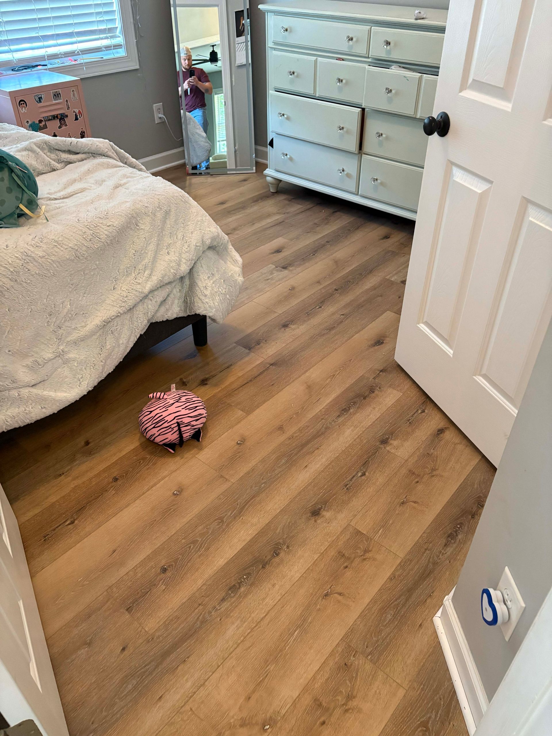 Bedroom with wood-look flooring. A bed, dresser, and open doorway are visible. A small child stands in the doorway.