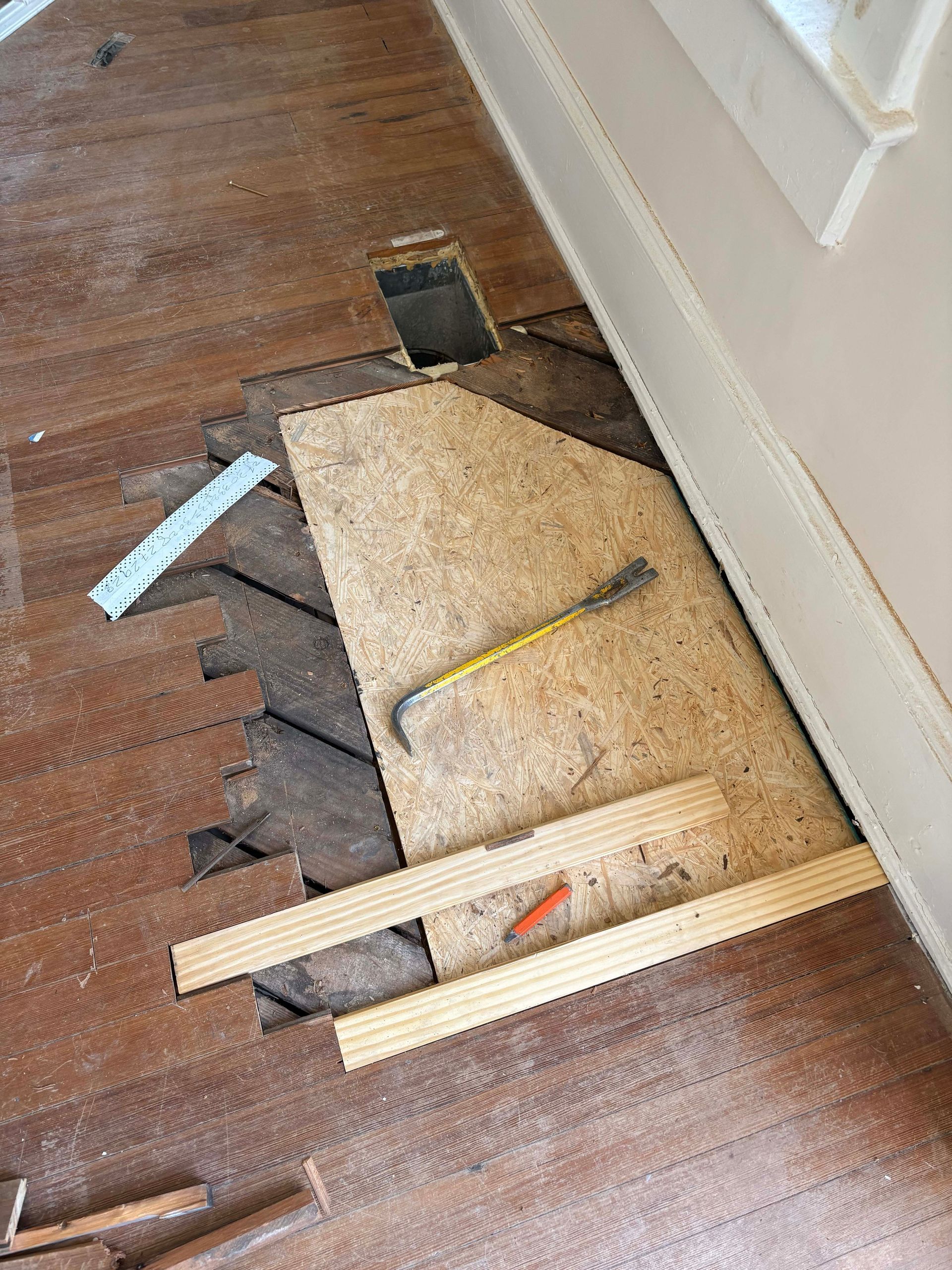 Floorboards being removed during renovation, revealing subfloor and a vent. A pry bar, and boards are present.