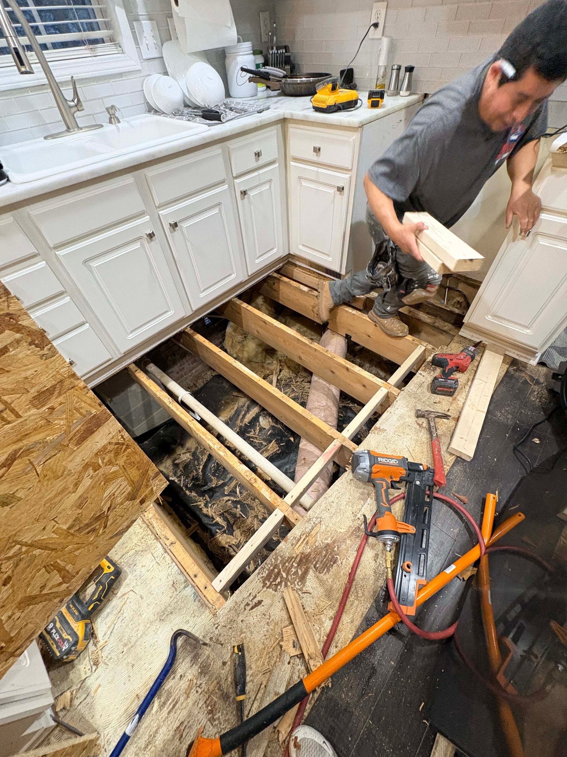 Man repairing a kitchen floor, which is open to the subfloor. Cabinets and appliances are present.