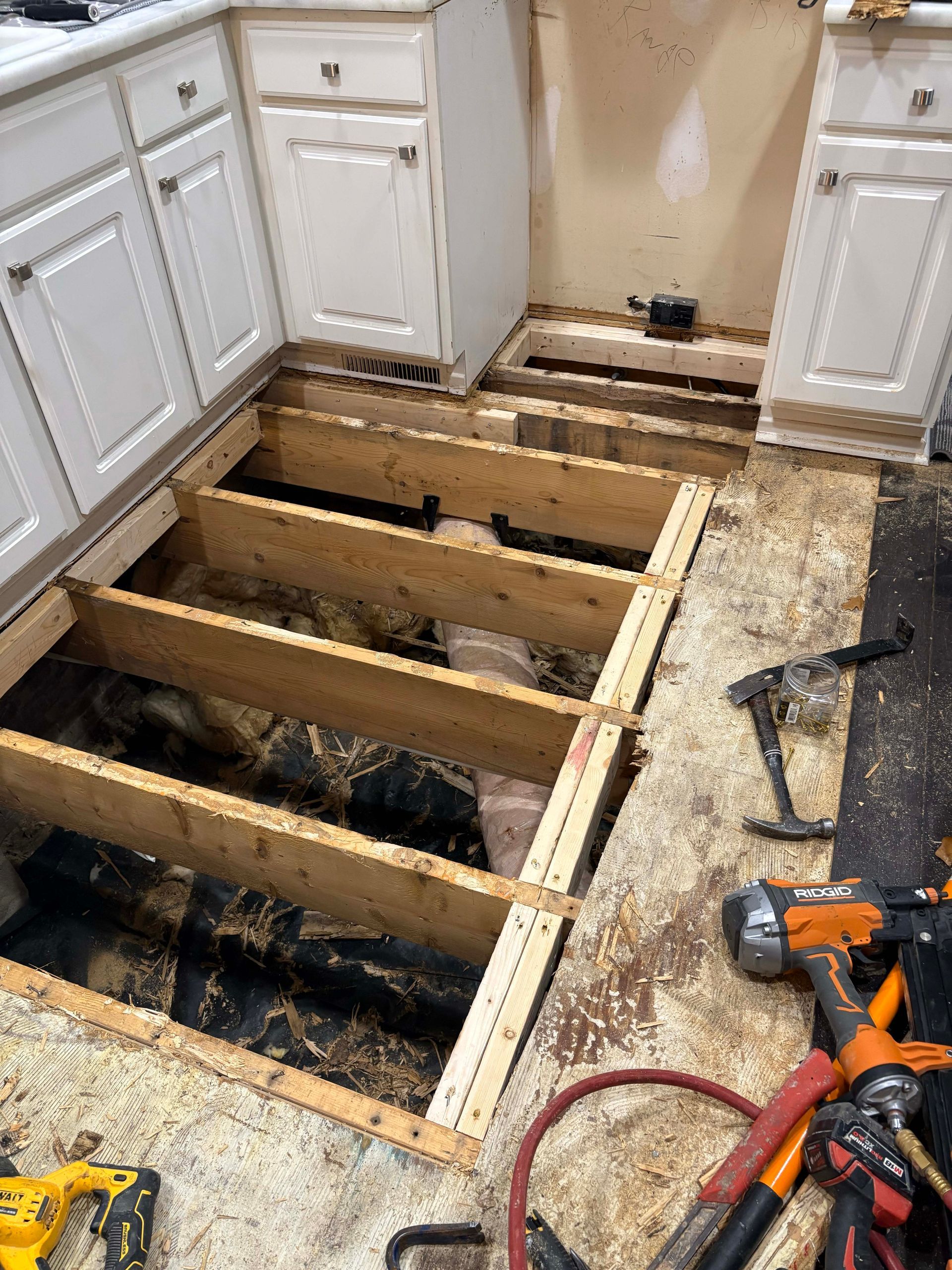 Kitchen floor under construction, exposing joists and subfloor. Cabinets visible, tools present.