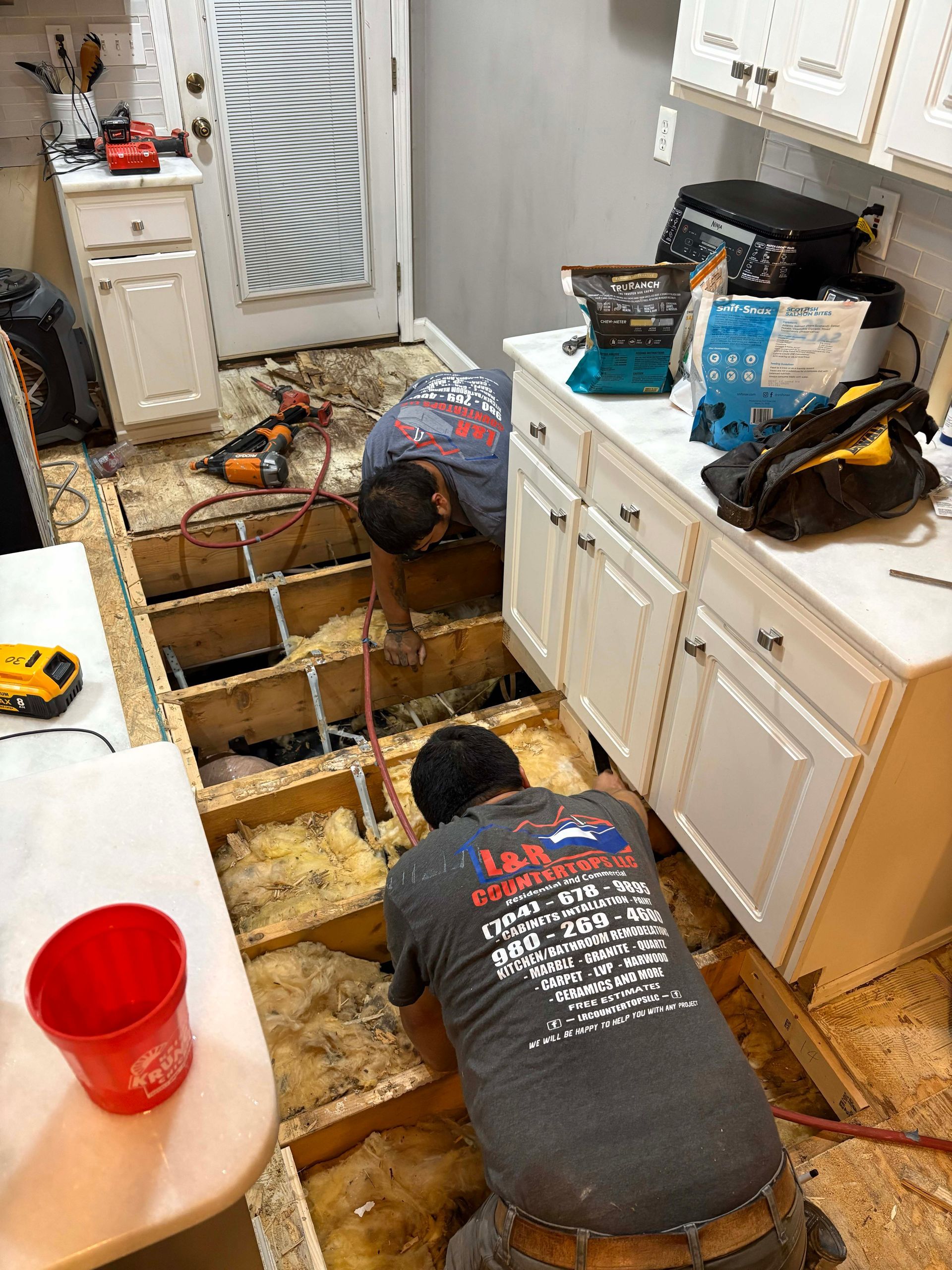 Two workers repairing a kitchen floor, one using tools, with exposed subfloor and insulation.