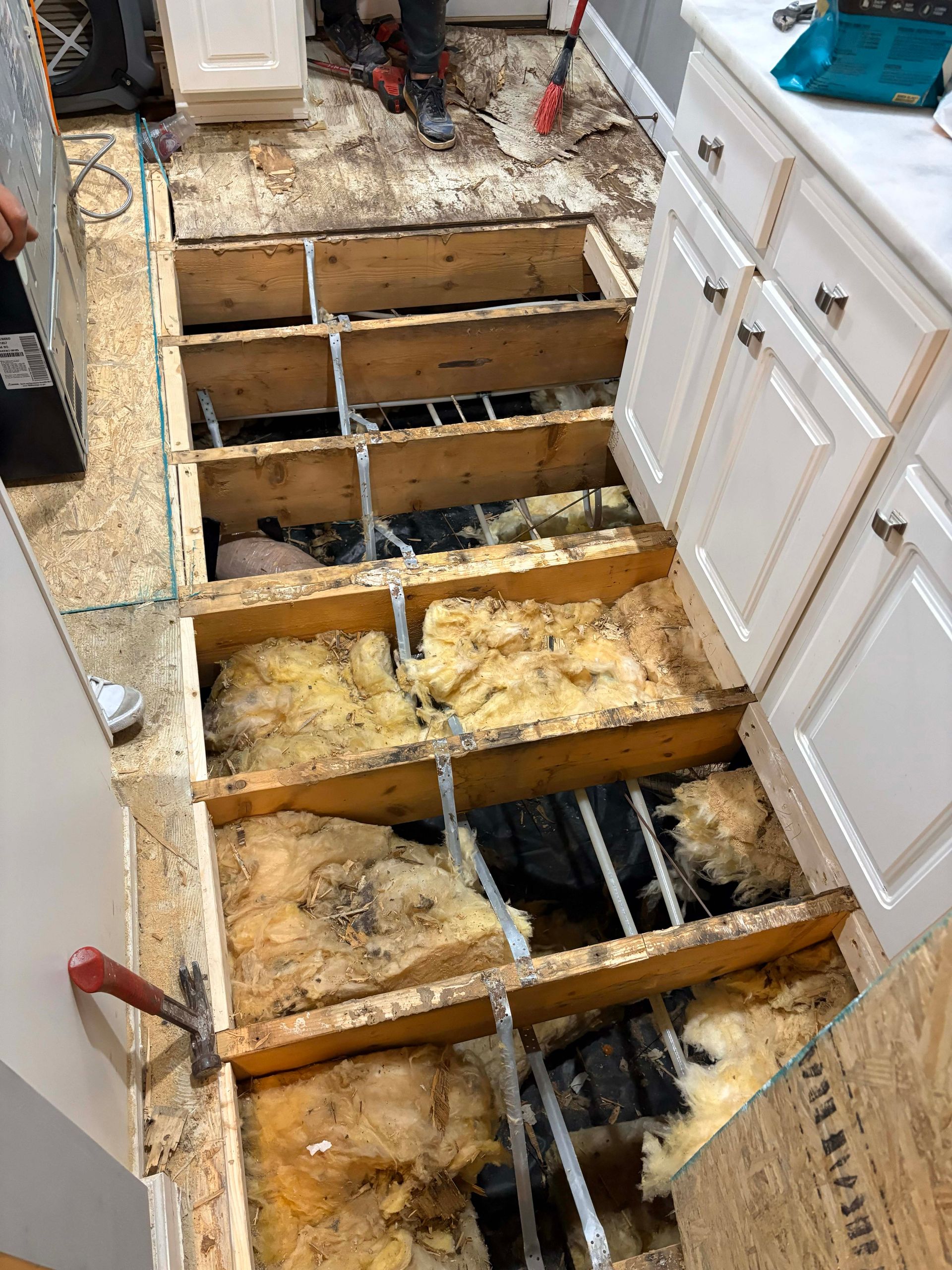 Kitchen floor under construction, exposed joists, insulation, and subfloor removed. Hammer and cabinets visible.
