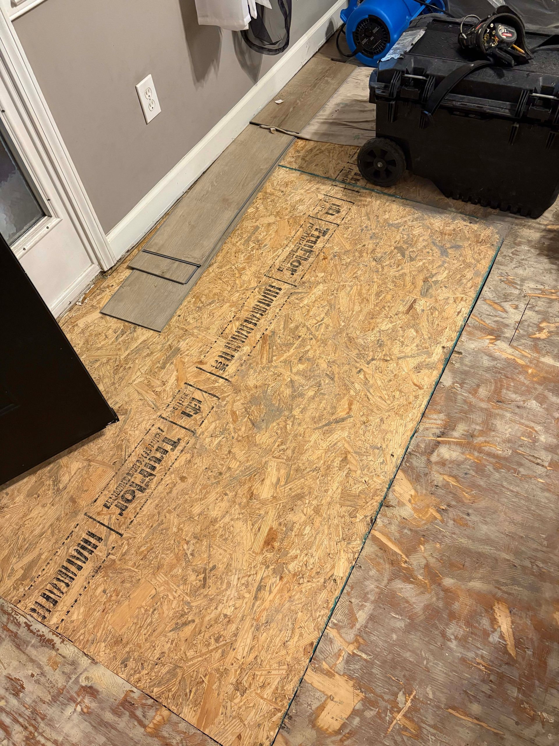Damaged flooring repair: plywood patch on damaged subfloor, door frame, and drying equipment visible.