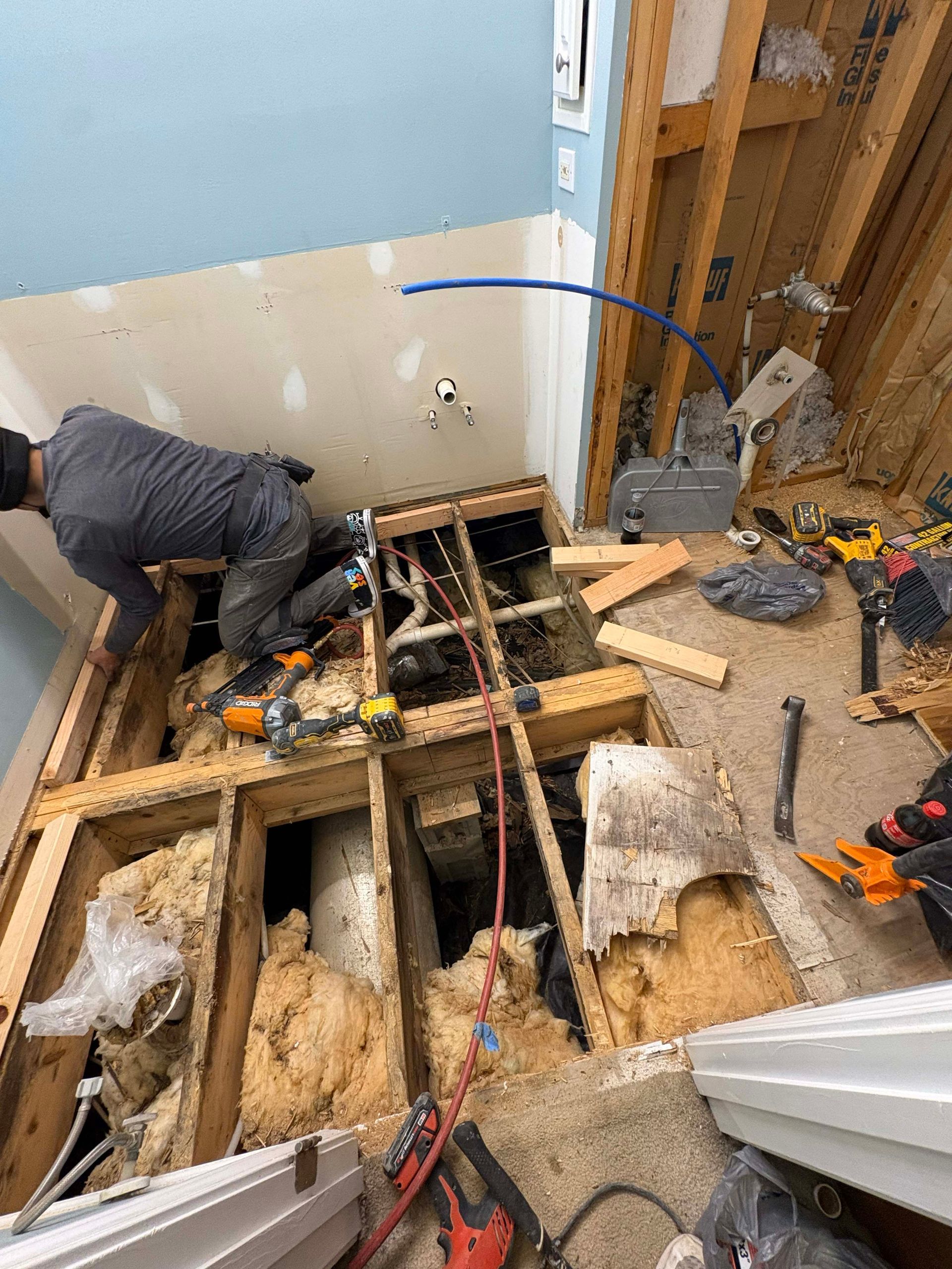 A person works on exposed plumbing in a bathroom with damaged flooring and insulation; tools and debris are scattered.