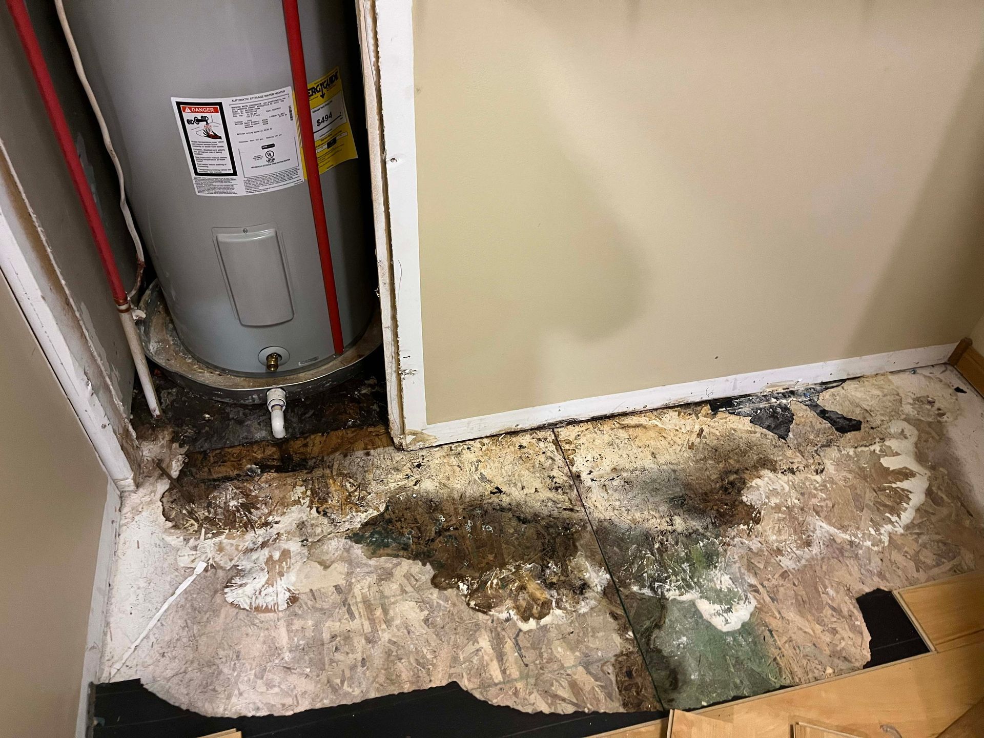Water heater in small alcove with damaged flooring. Brown and white water damage visible.
