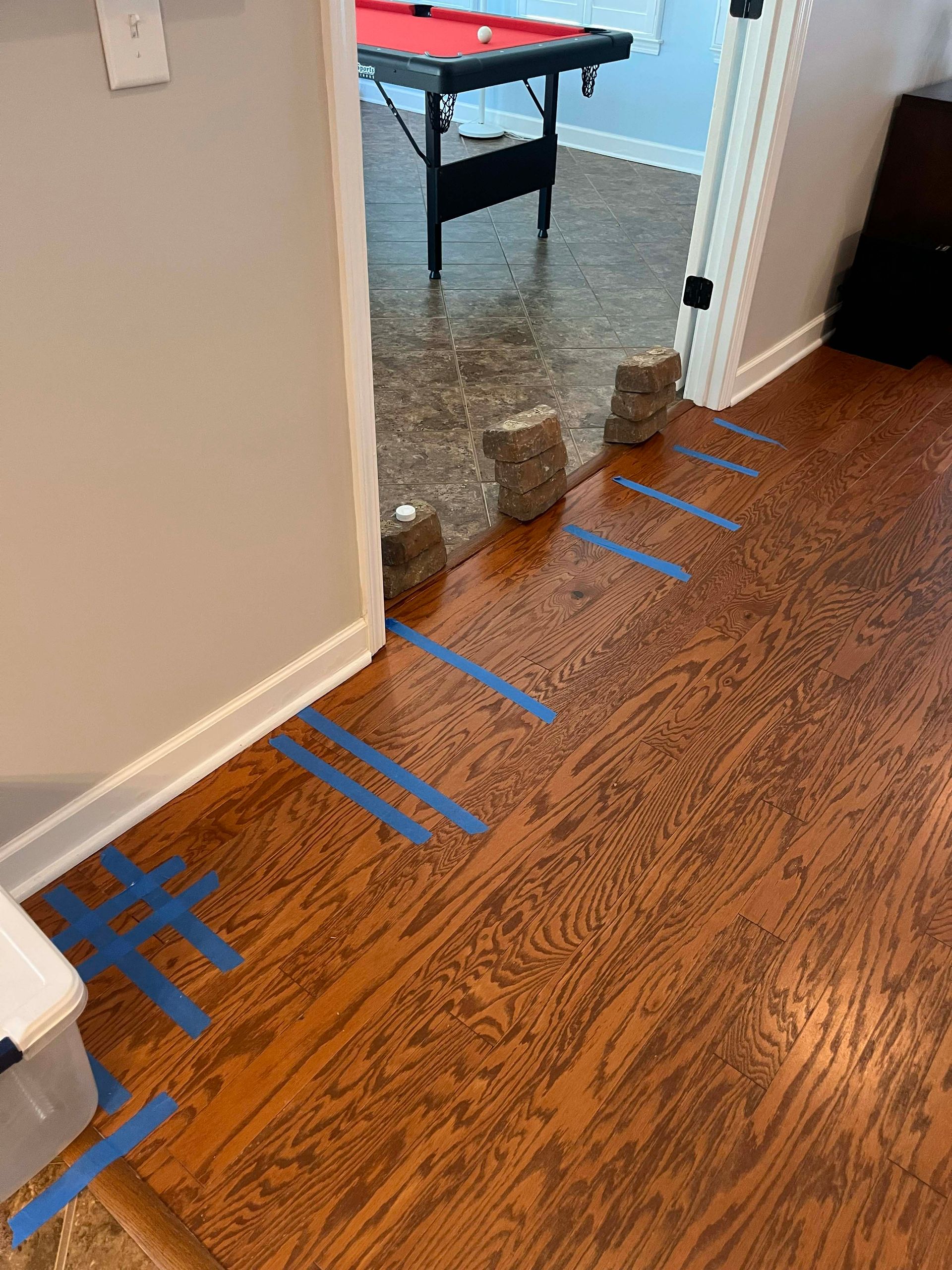 Hardwood floor with blue tape lines and stacked bricks blocking a doorway to a pool table room.