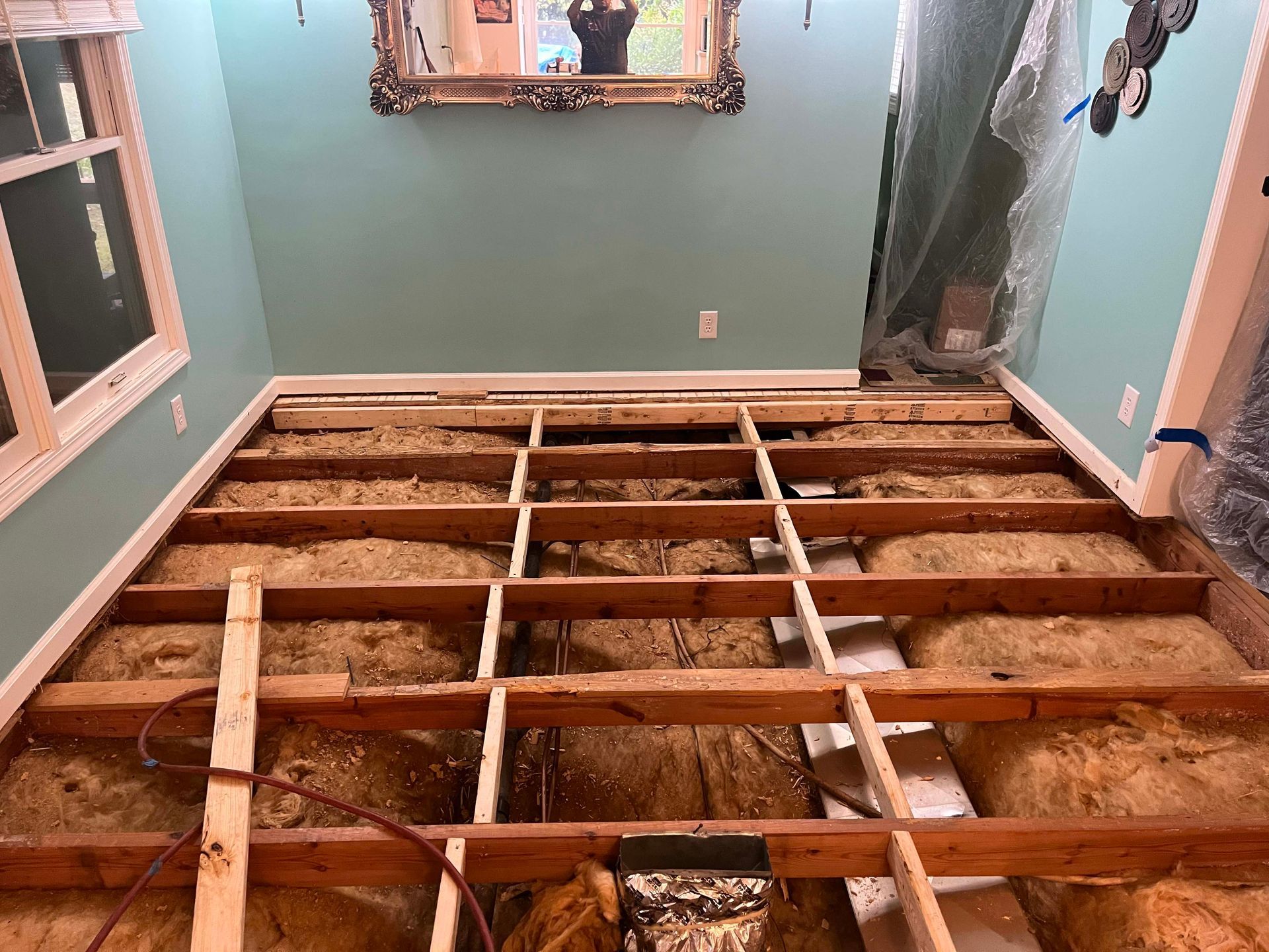 Room with exposed floor joists, insulation, and partially demolished floor. Teal walls, a window, and a mirror are visible.