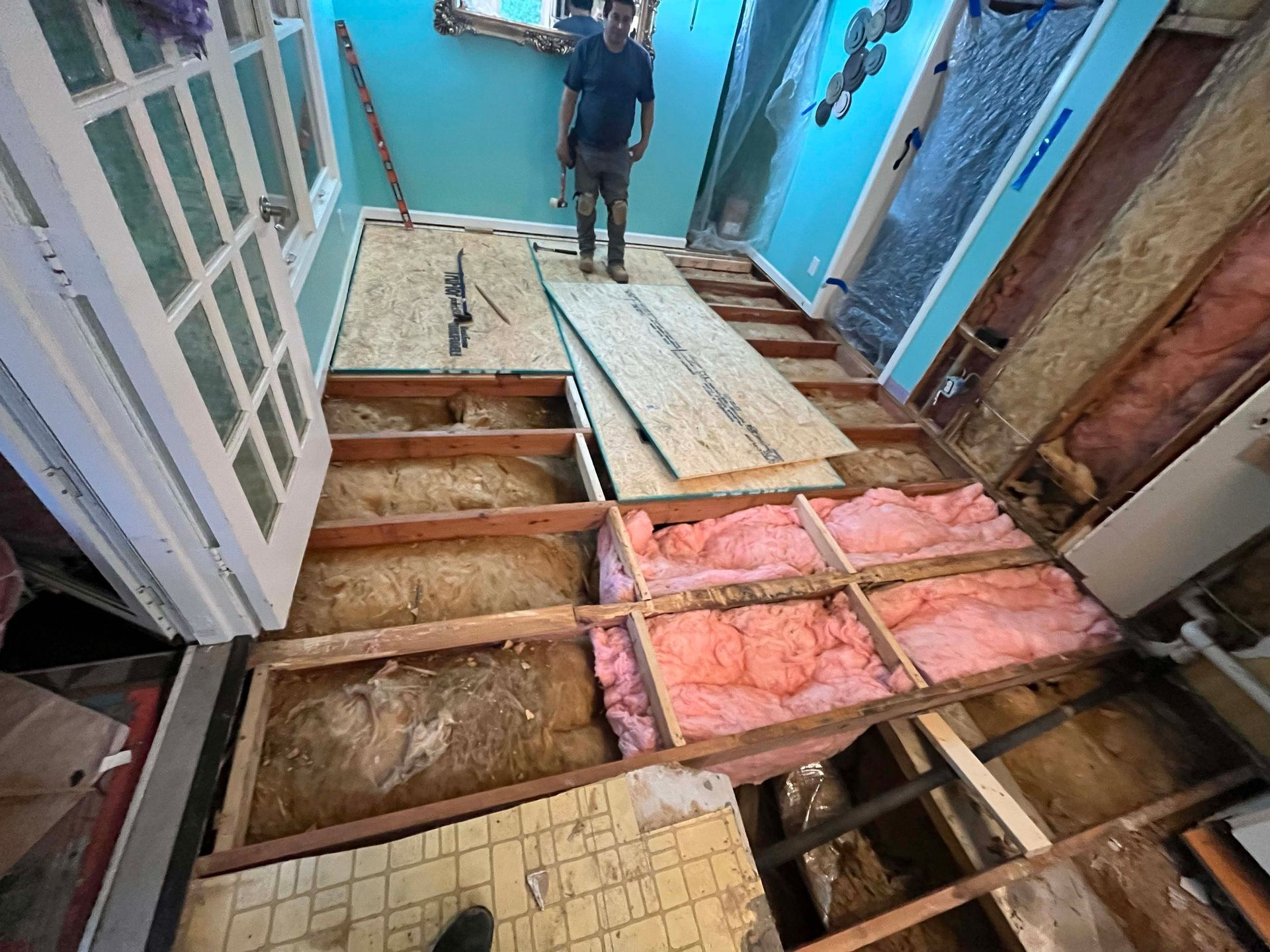 Man standing in room with exposed floorboards and insulation during renovation.