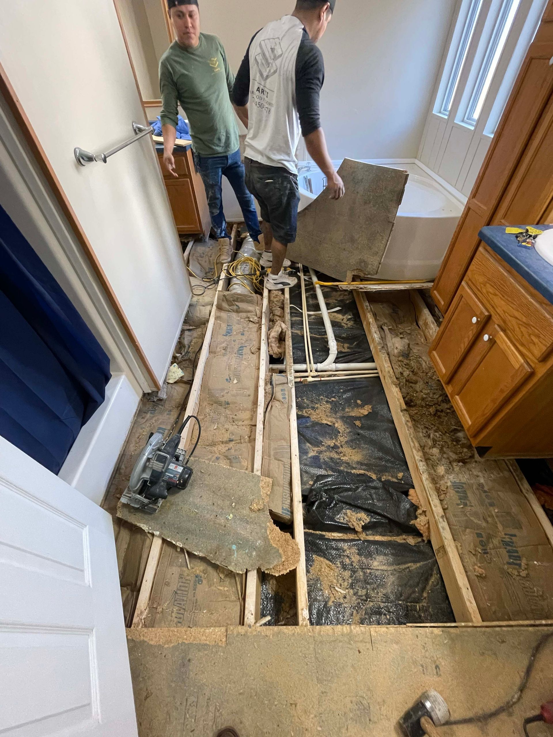 Two men renovating a bathroom, removing floorboards, water pipes visible.