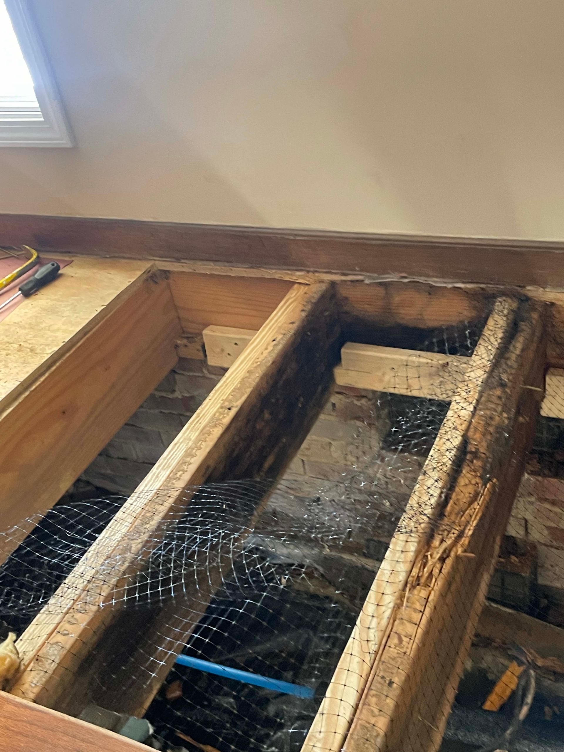 Open floor joists with black mesh and blue pipe visible, likely under renovation.
