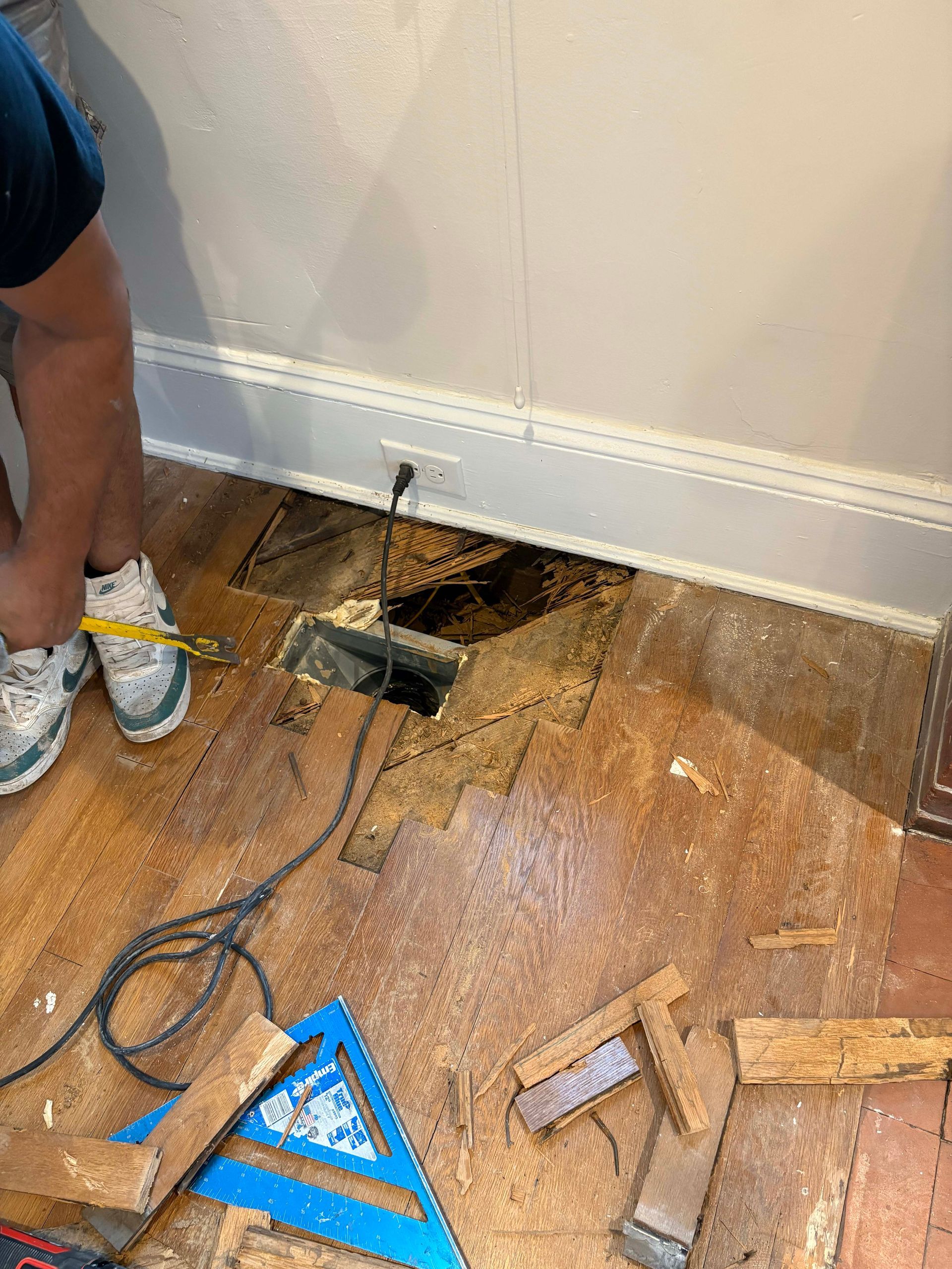 Person working on a damaged wood floor near a wall with a vent.