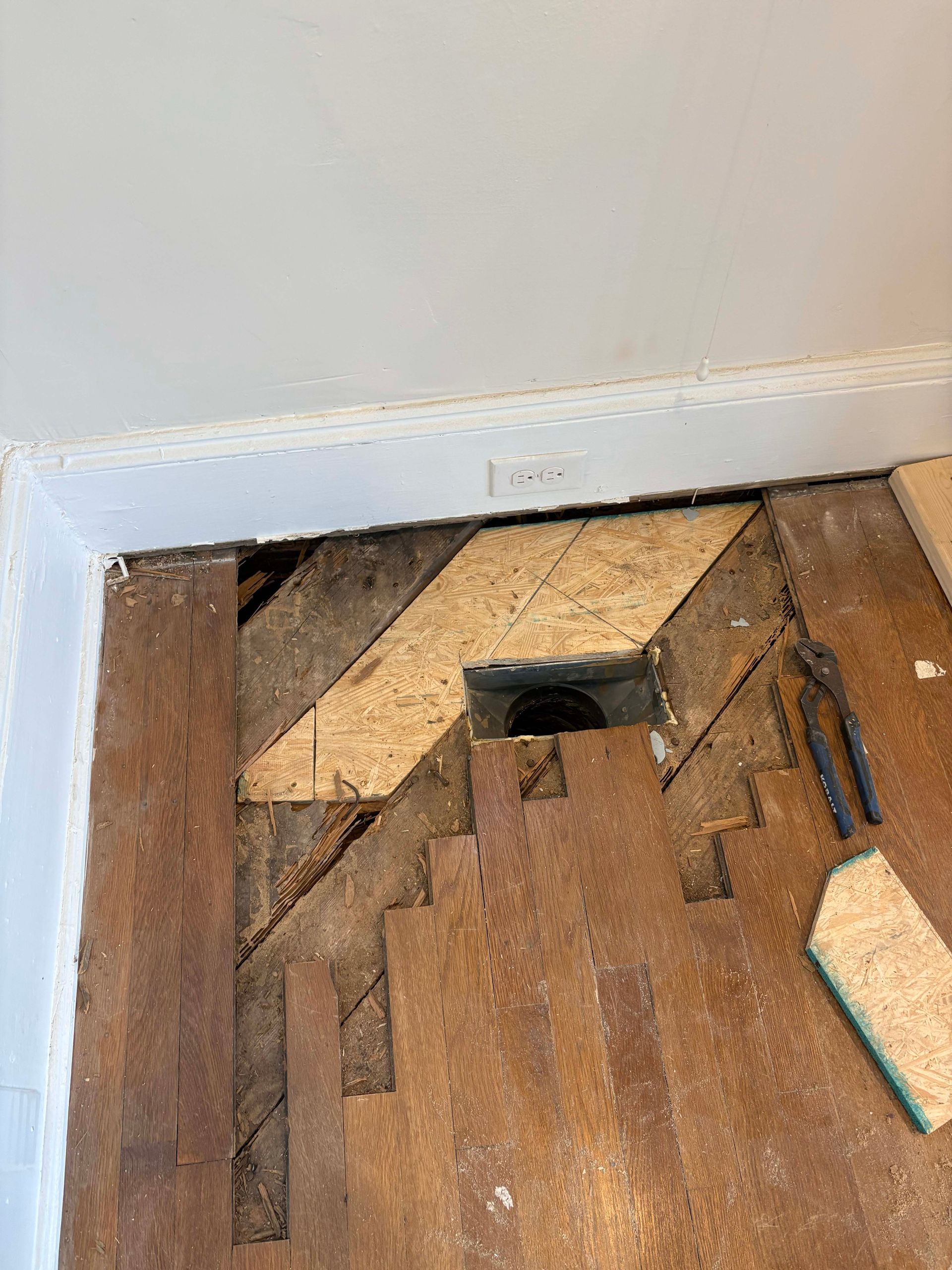 Floor repair: Exposed subfloor and damaged hardwood around a plumbing access point.