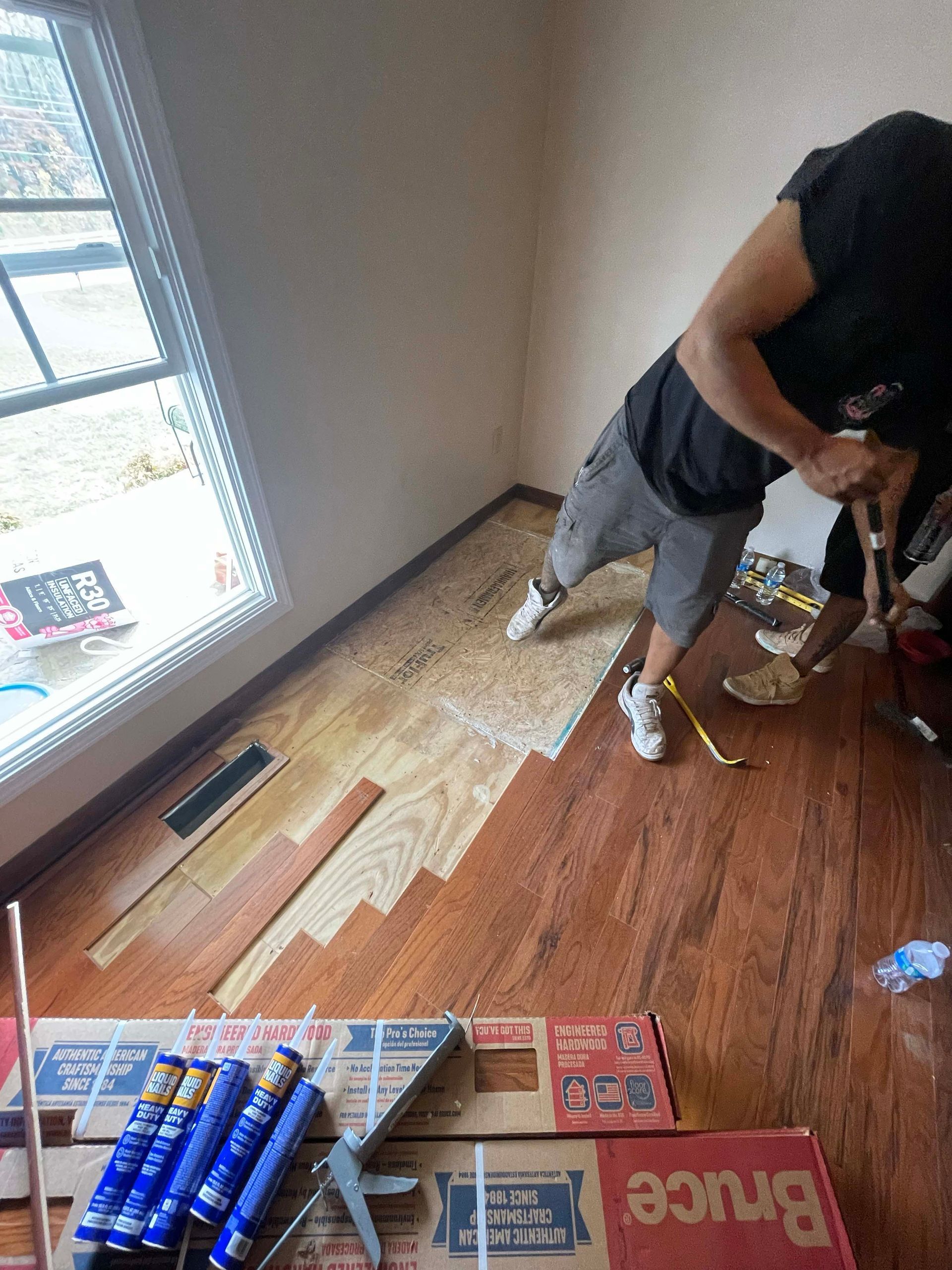 Man installing hardwood flooring near a window. He uses tools, brown wood, in a room.