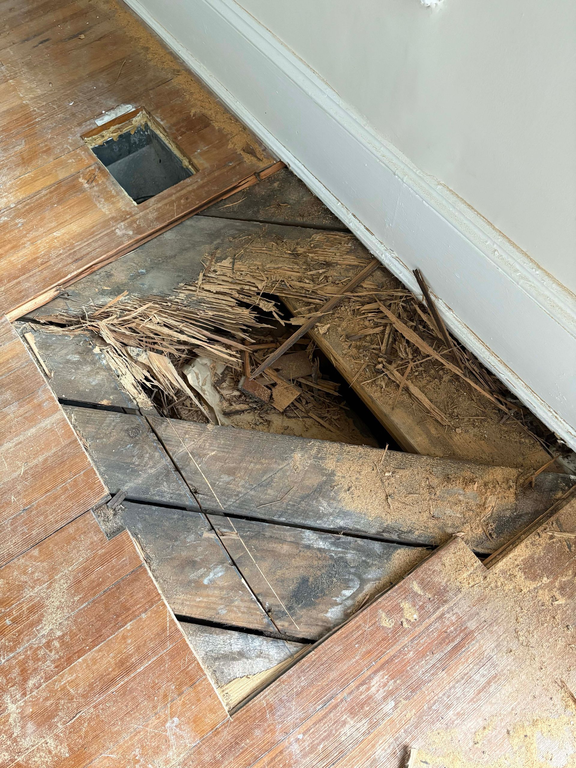 Damaged wooden floor with a large rectangular hole, revealing subfloor and debris.
