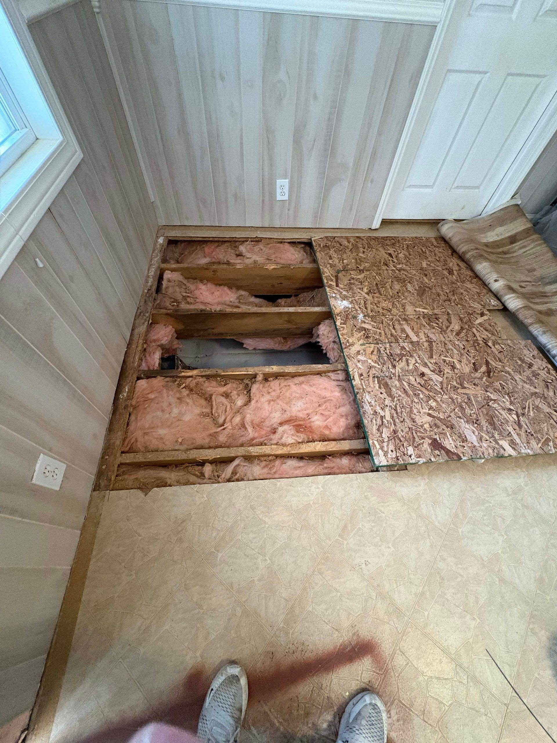 Damaged flooring exposing wood beams and insulation. A person’s feet in sneakers are visible.