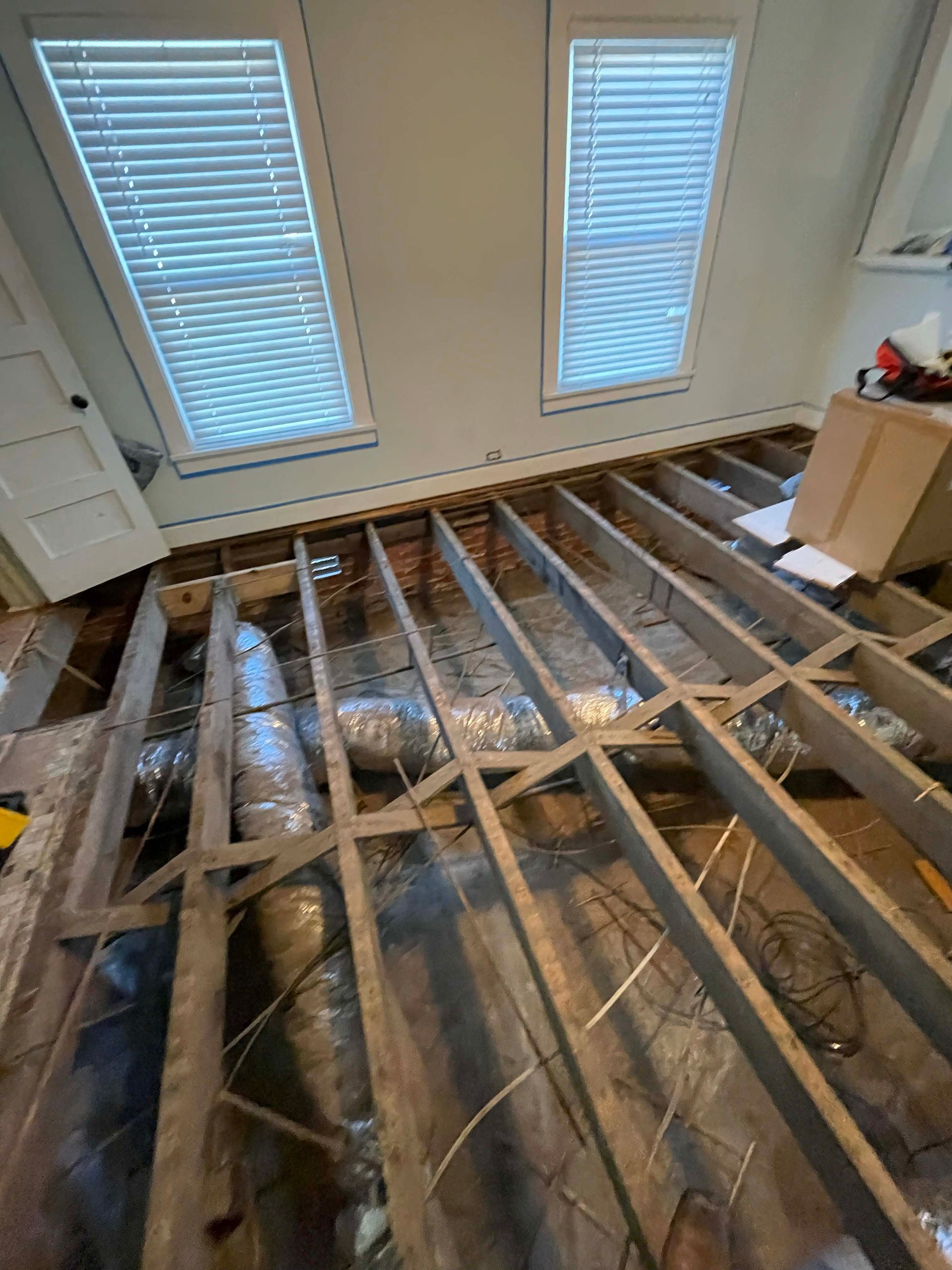 Room with floorboards removed, revealing joists and insulation beneath two windows.