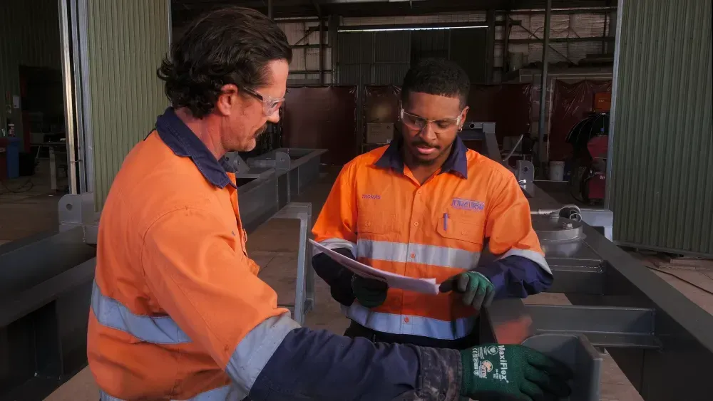 Two Workers in Orange Workwear Reviewing Paperwork in a Workshop — Junction Engineering Group in Evans Landing, QLD