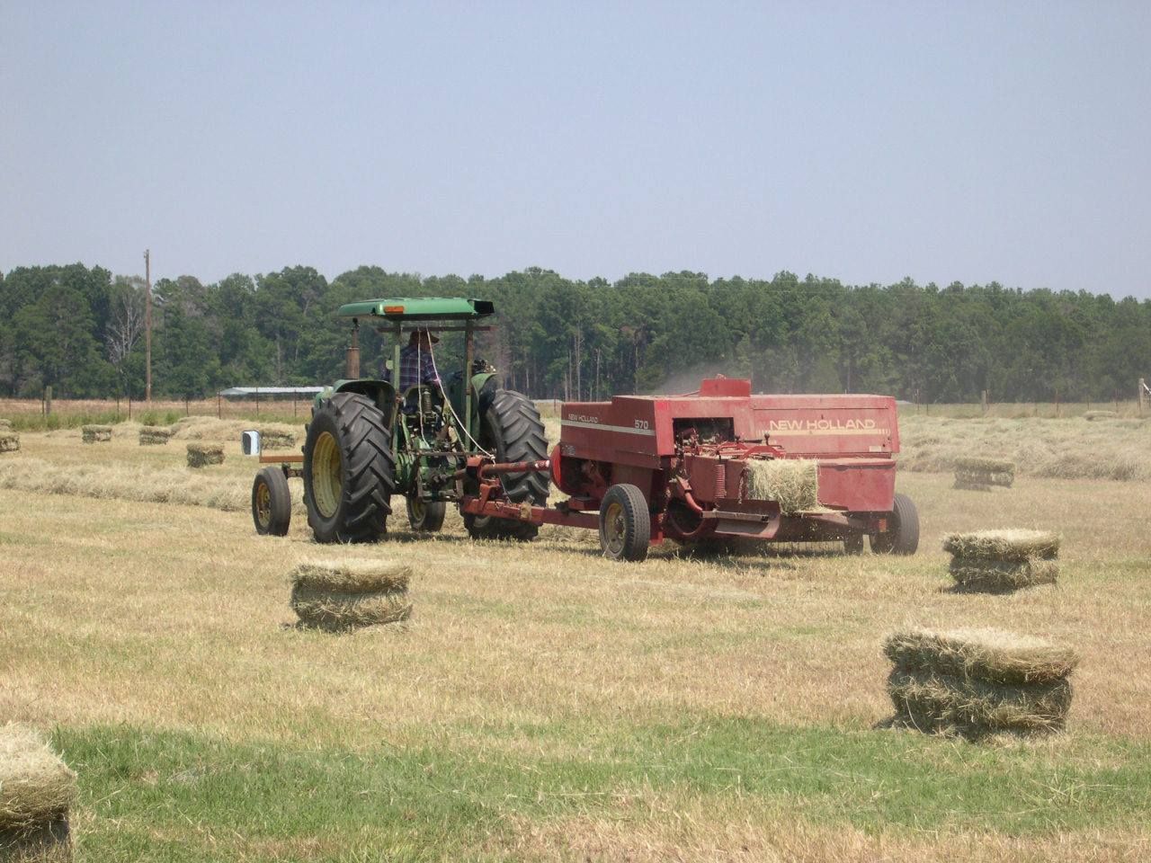 Tractor hauling hay baler in a field with several hay bales and a tree line in the background